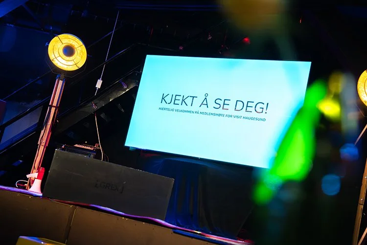 Et sceneteppe med en stor skjerm som viser teksten "KJEKT Å SE DEG!" og en mindre tekst under som sier "Hjertelig velkommen på medlemsmøte for Visit Haugesund". Scenen har belysning og utstyr for en presentasjon.