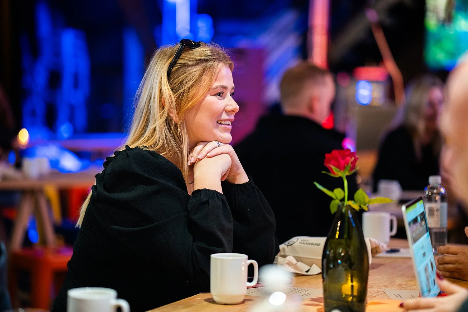 En kvinne med blondt hår smiler mens hun sitter ved et bord i en ufullstendig utescene med flere personer i bakgrunnen. På bordet er det en rød rose i en vase, kopper, en vannflaske og en mobiltelefon.