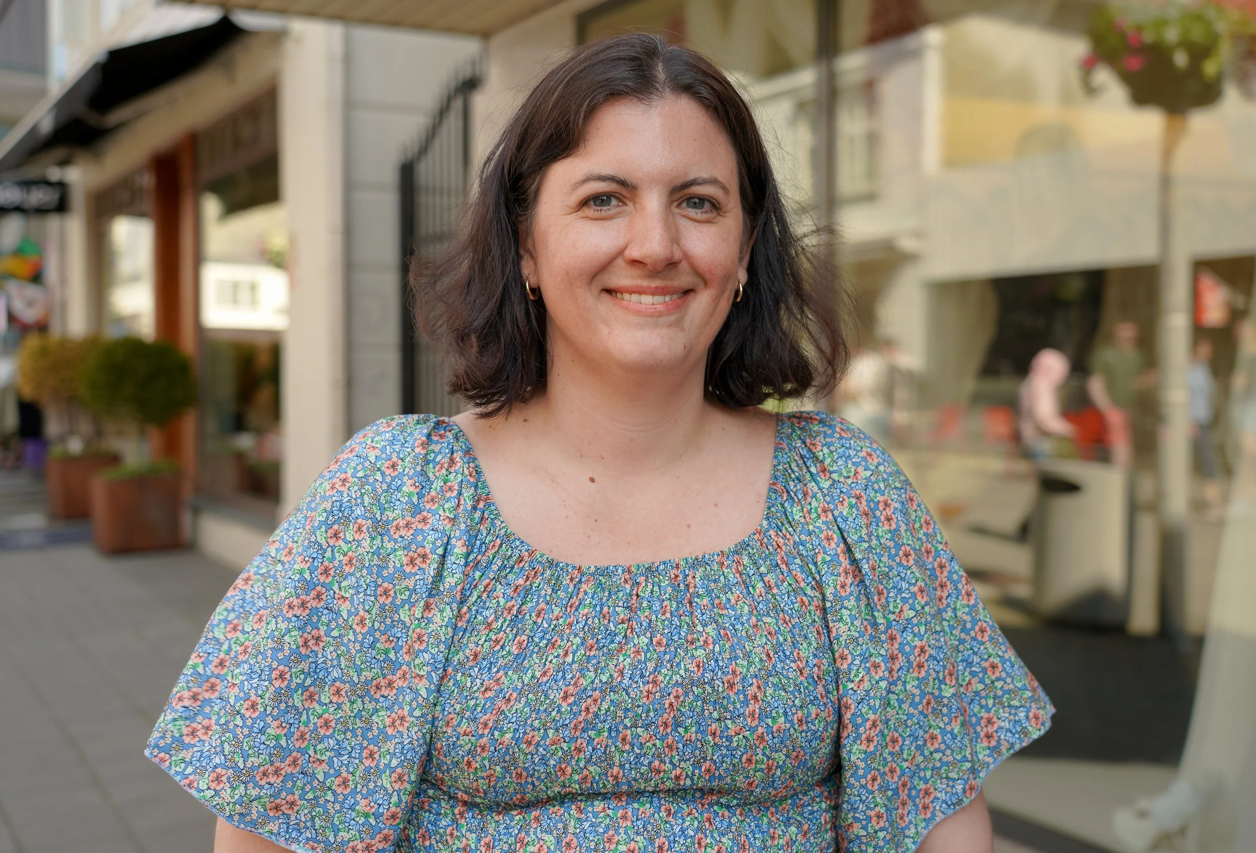 Et portrett av en smilende kvinne med mørkt, skulderlangt hår, iført en blå blomstret topp, som står ute på gaten med butikker og mennesker i bakgrunnen.