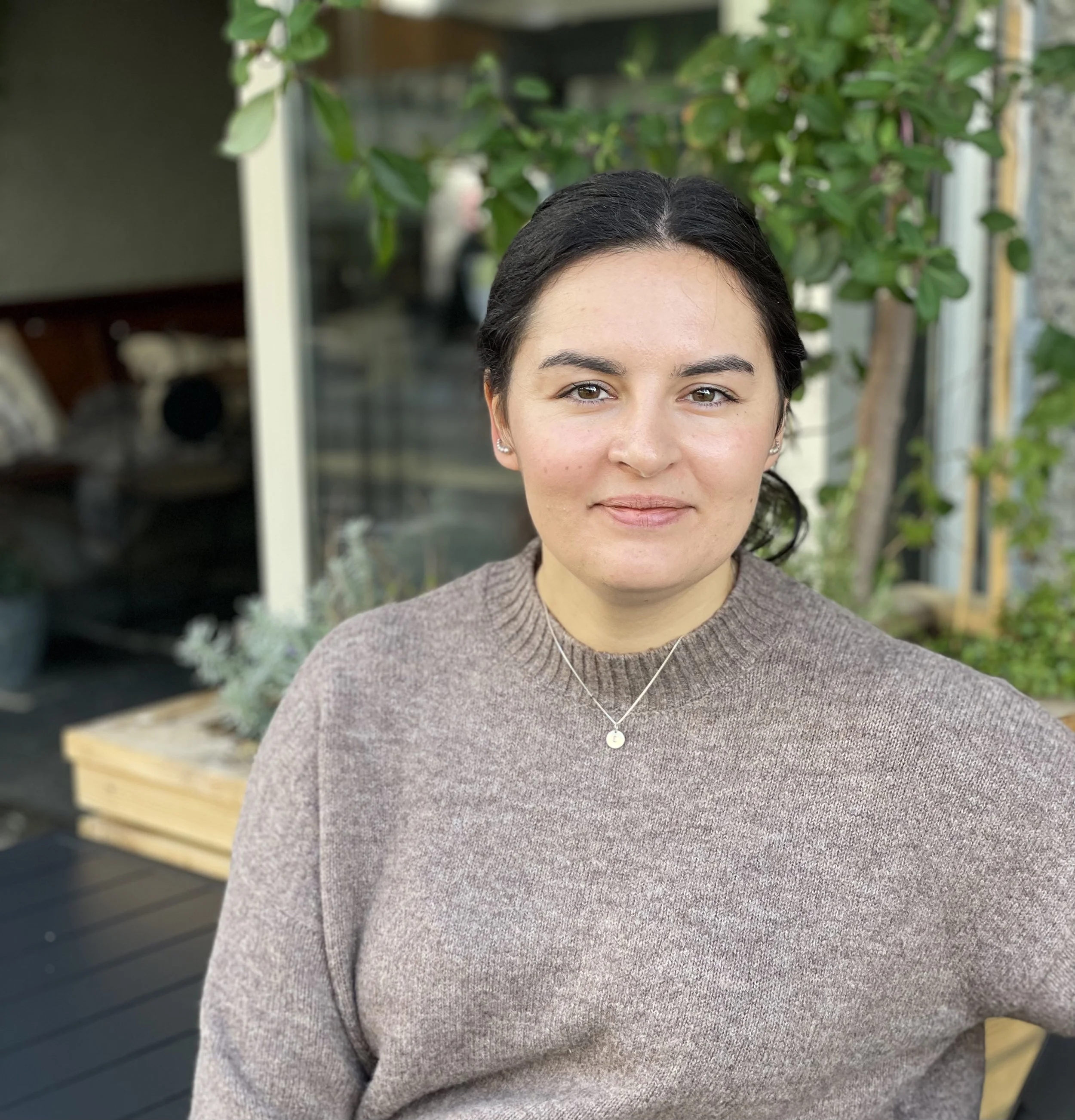 En kvinne i en grå genser, sitter utendørs foran et vindu med grønt løvverk i bakgrunnen.