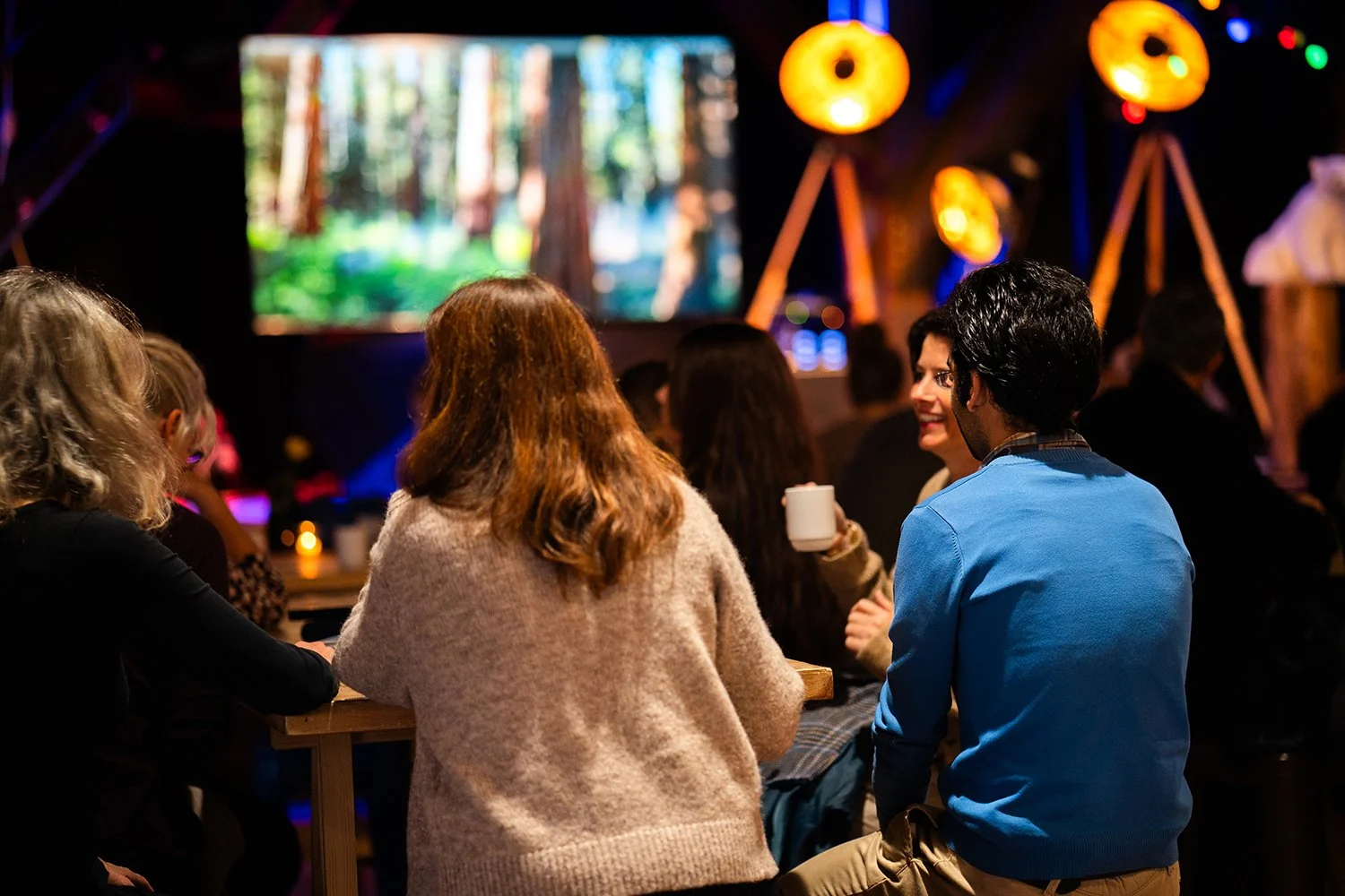 Folk som sitter og snakker i et moro bar- eller kafemiljø med lysende lamper og et stort lerret som viser en natur- eller skogscene.