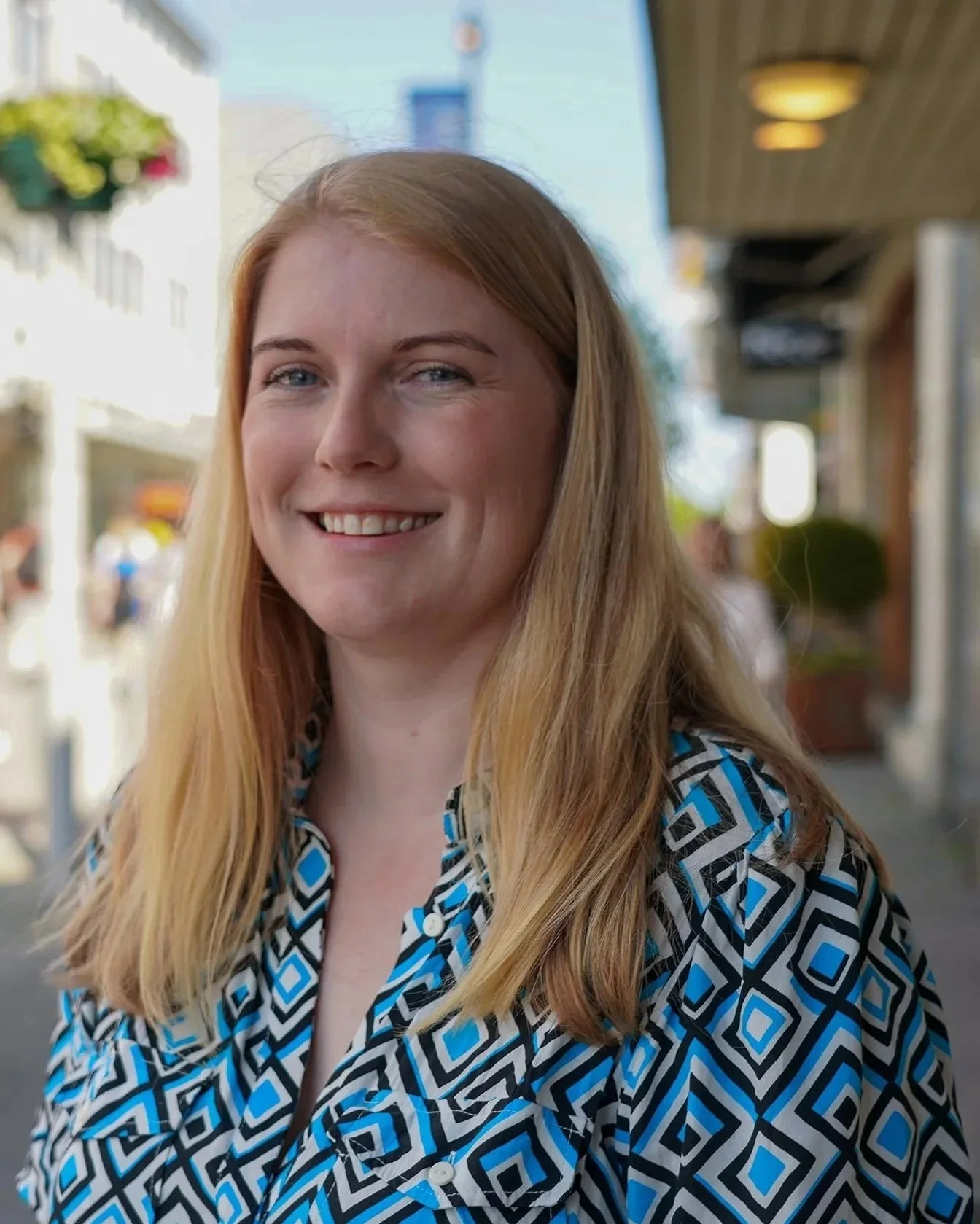 Ung kvinne med lang, blond hår smilende på gaten i solskinn, iført en blå, svart og hvit mønstret skjorte.