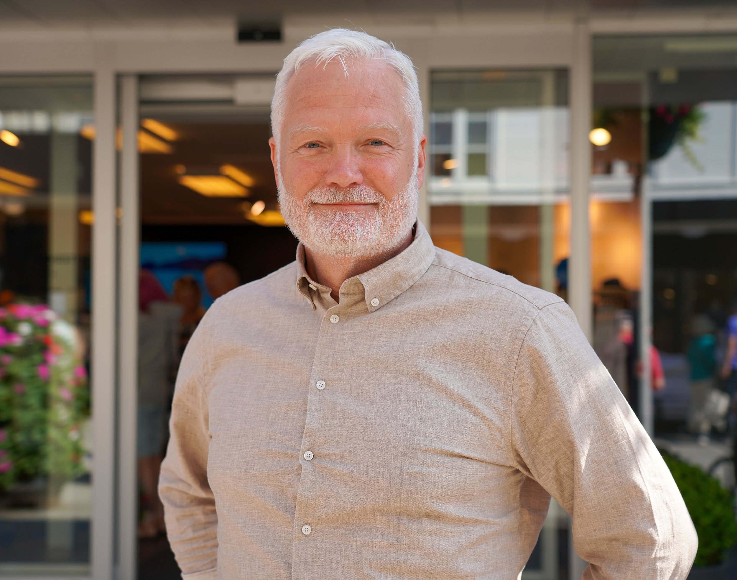 En eldre man med hvit hår og skjegg, smilende, står foran en bygning med glassdører, iført en lys brun skjorte. Bakgrunnen viser et urbant miljø med mennesker og blomster.