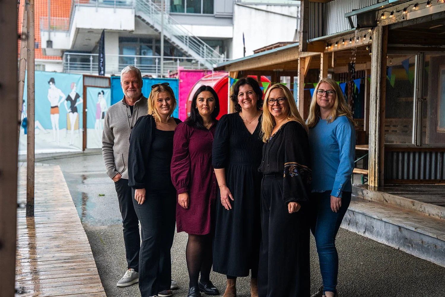 Seks personer står utenfor, smilende, med bakgrunn av en vegg med bilder og en tilfeldig bygning, under vannet flagg og dekorasjoner.