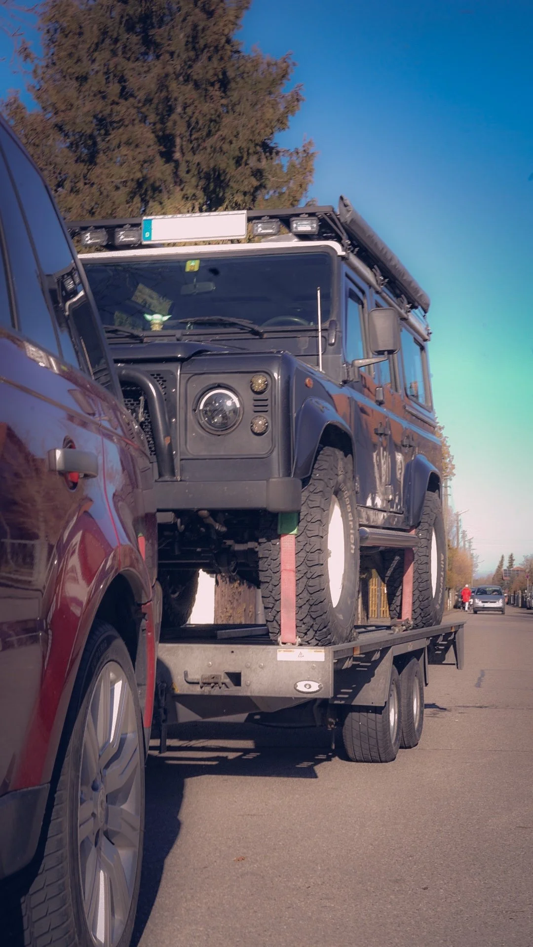 A black Land Rover Defender loaded on a flatbed tow truck, with a tree and a few cars visible in the background on a clear day.