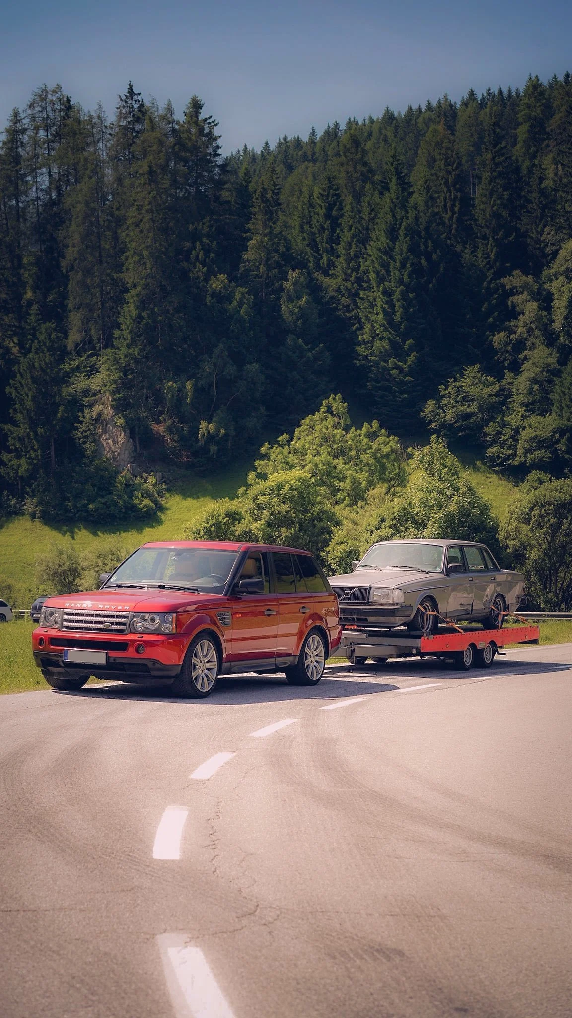 A red Range Rover towing a vintage gray car on a trailer, parked on a curve of a paved road with green hills and dense trees in the background.
