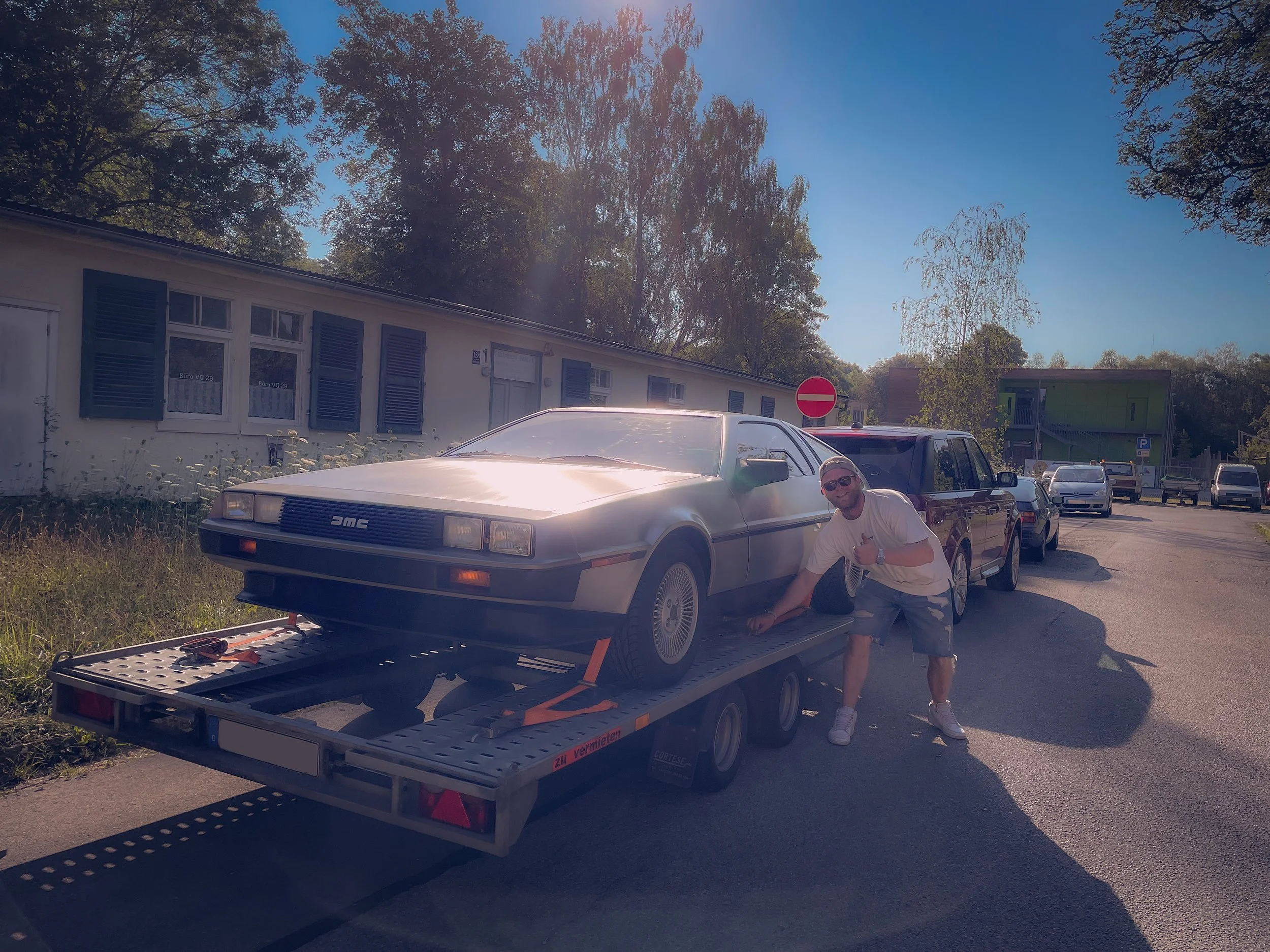 A man wearing sunglasses and a white shirt is kneeling beside a DeLorean DMC car on a trailer. The trailer is parked on the side of a street with trees and buildings in the background. There is a no entry sign and several parked cars visible.