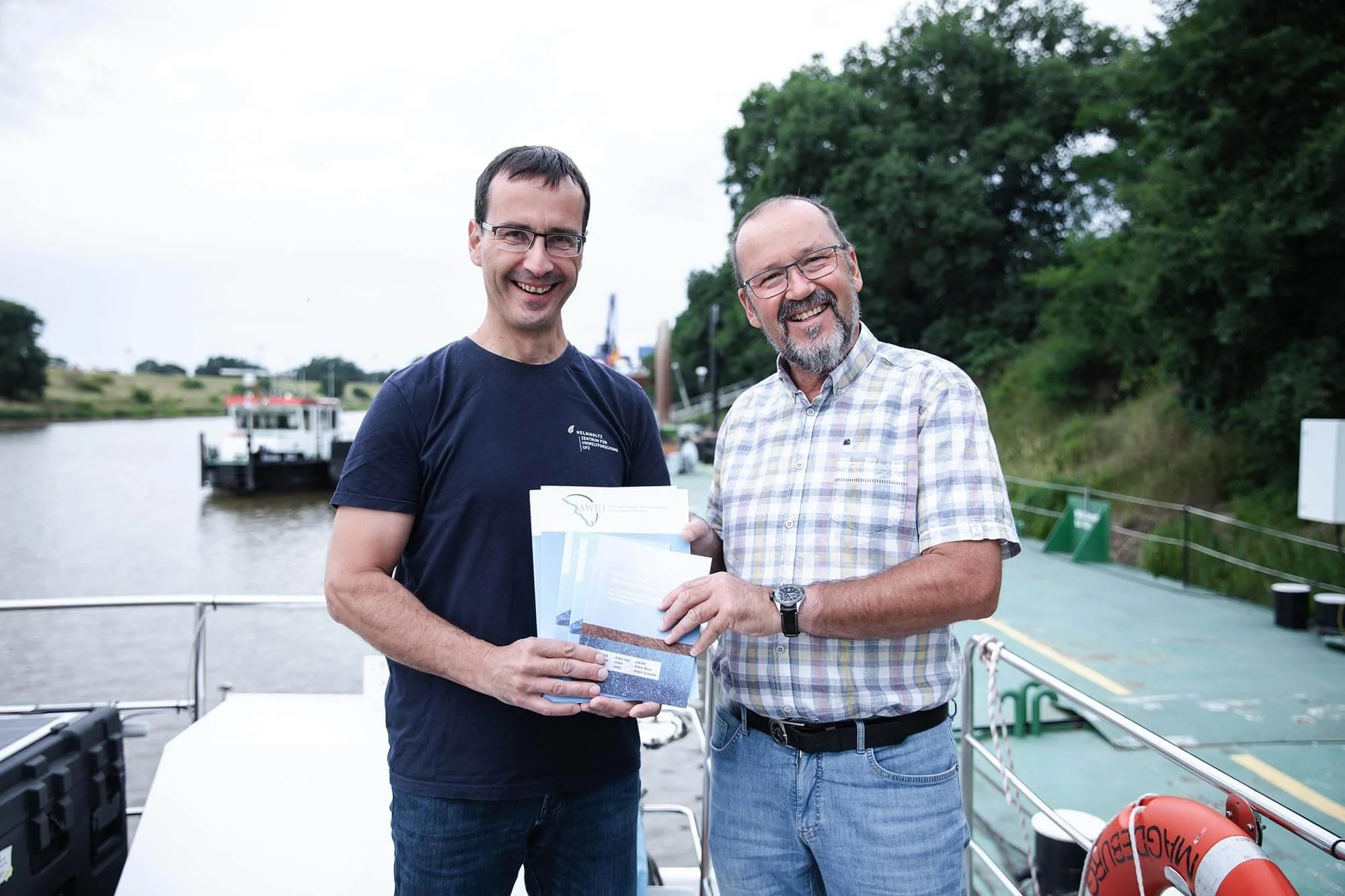 Zwei Männer halten einen Gütebericht zur Qualität von Fließgewässern in den Händen und schauen lächelnd in die Kamera.