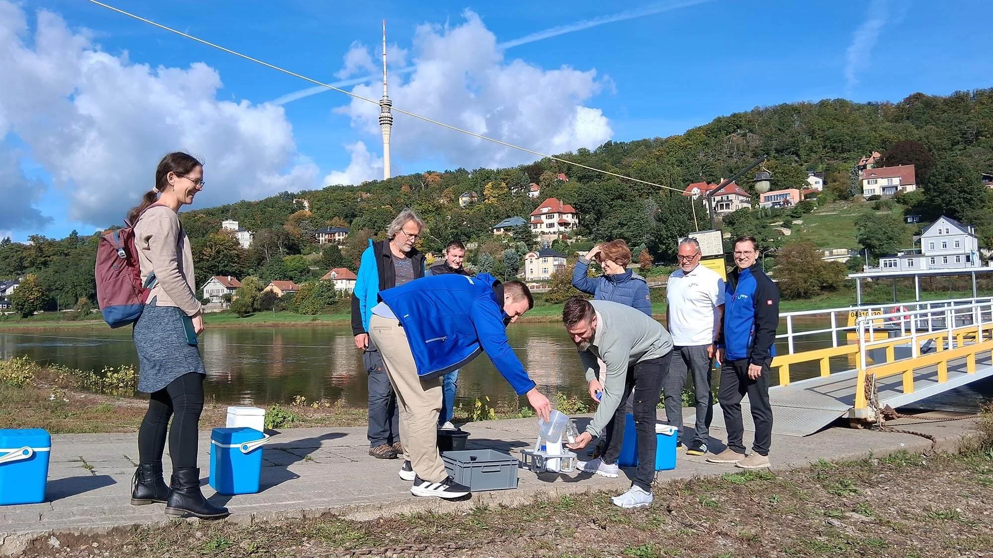 Eine Gruppe von Personen steht an der Elbe. Eine Person schenkt einer anderen Person Wasser aus einem Messbecher in ein Glas ein.