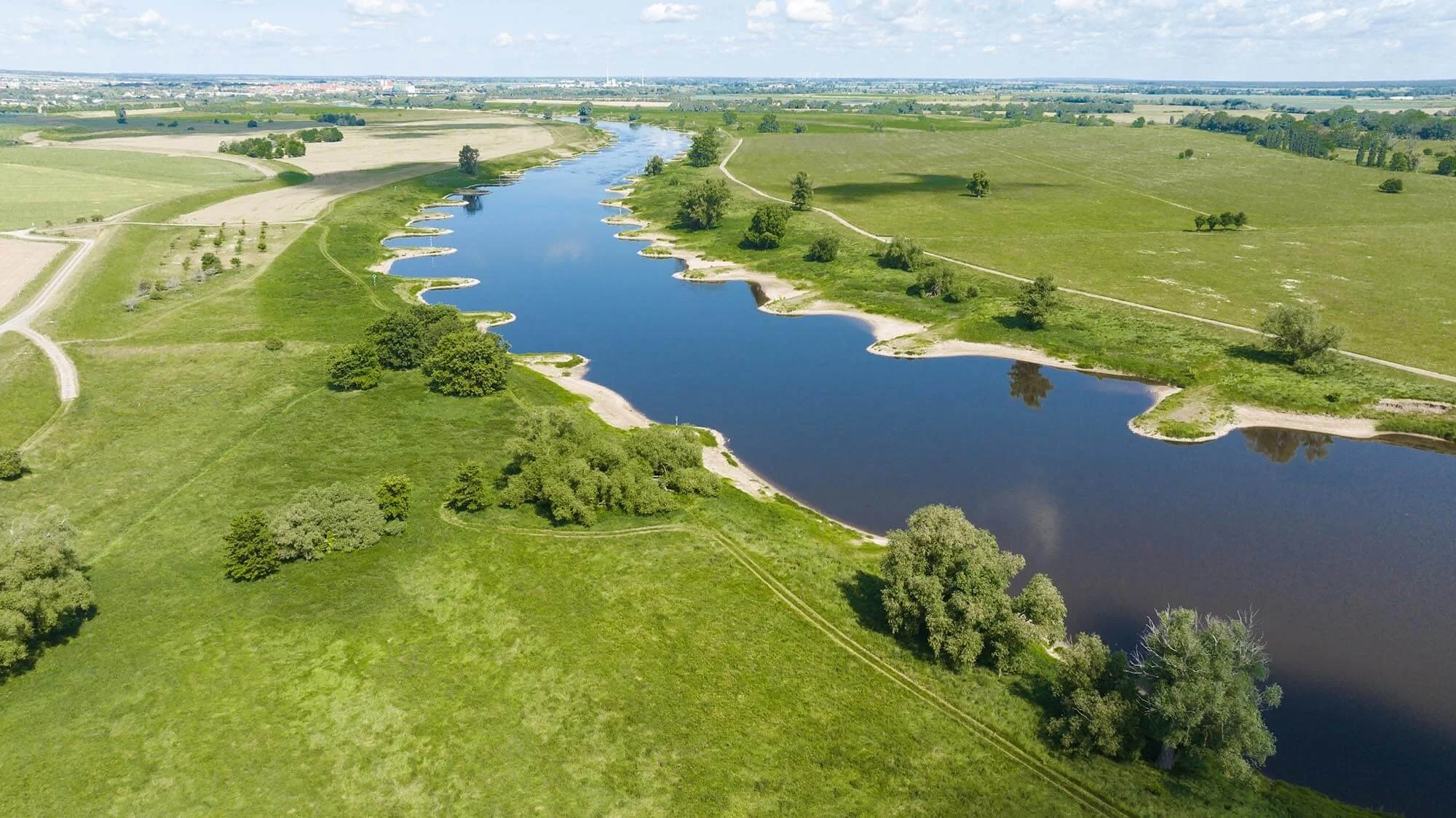 Der Fluss Elbe schlängelt sich durch eine grüne Aue mit Wiesen, Feldern und Bäumen. Man sieht ein kleines Stück Himmel.