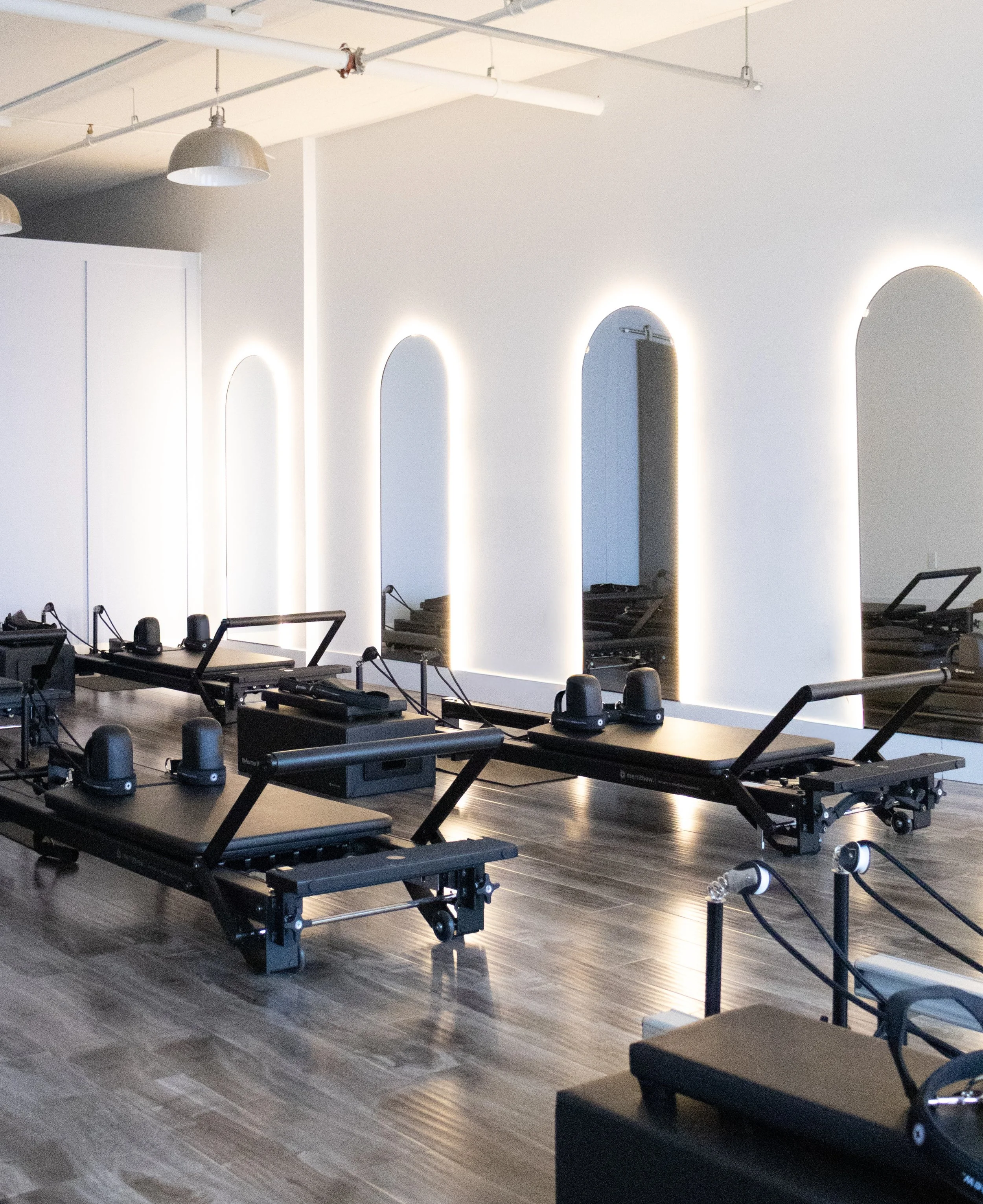 A Pilates studio with reformer machines arranged in rows, illuminated mirrors on the wall, and modern lighting fixtures.