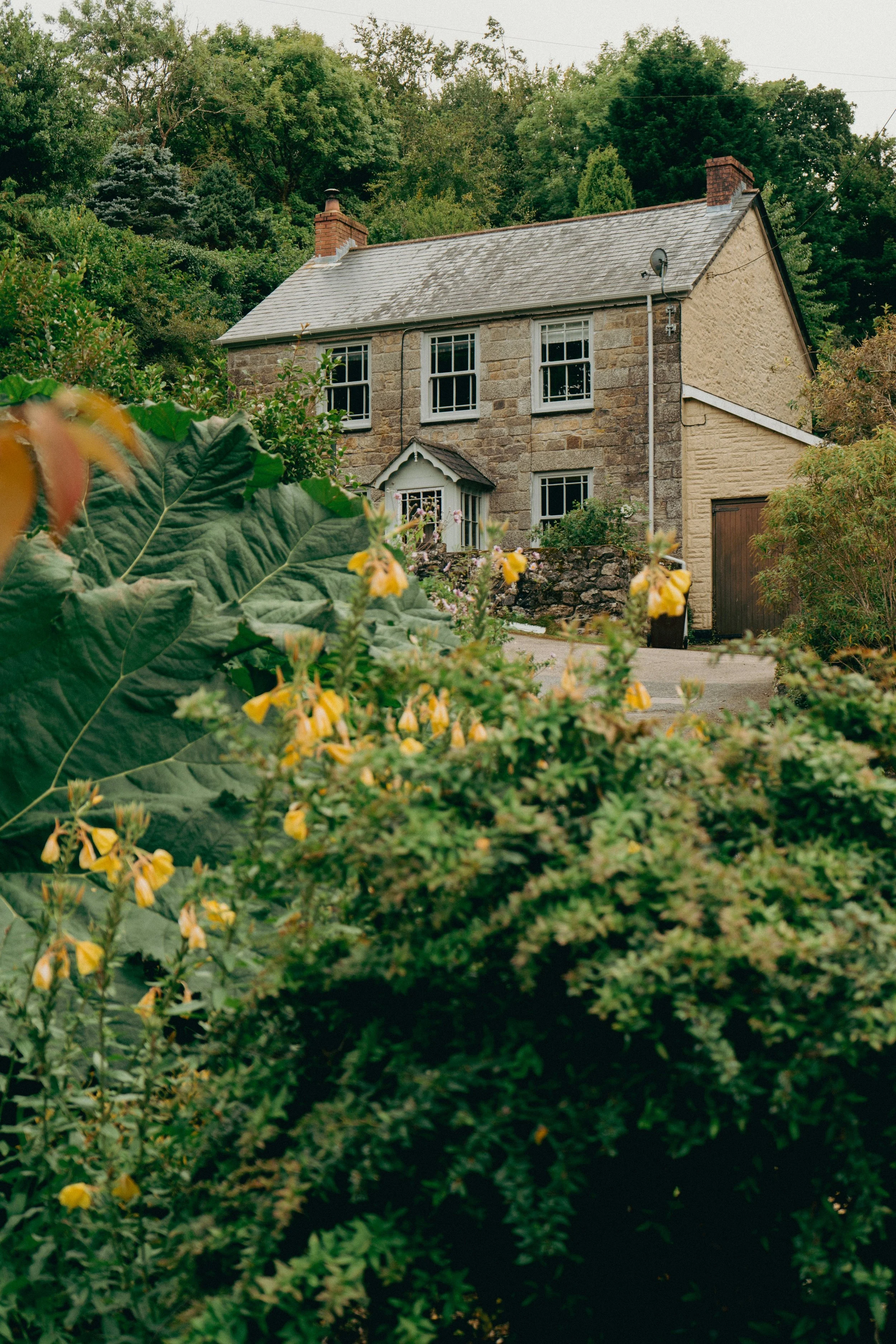A stone and brick house with a gray shingle roof, surrounded by lush green trees and shrubs, with yellow flowers in the foreground.