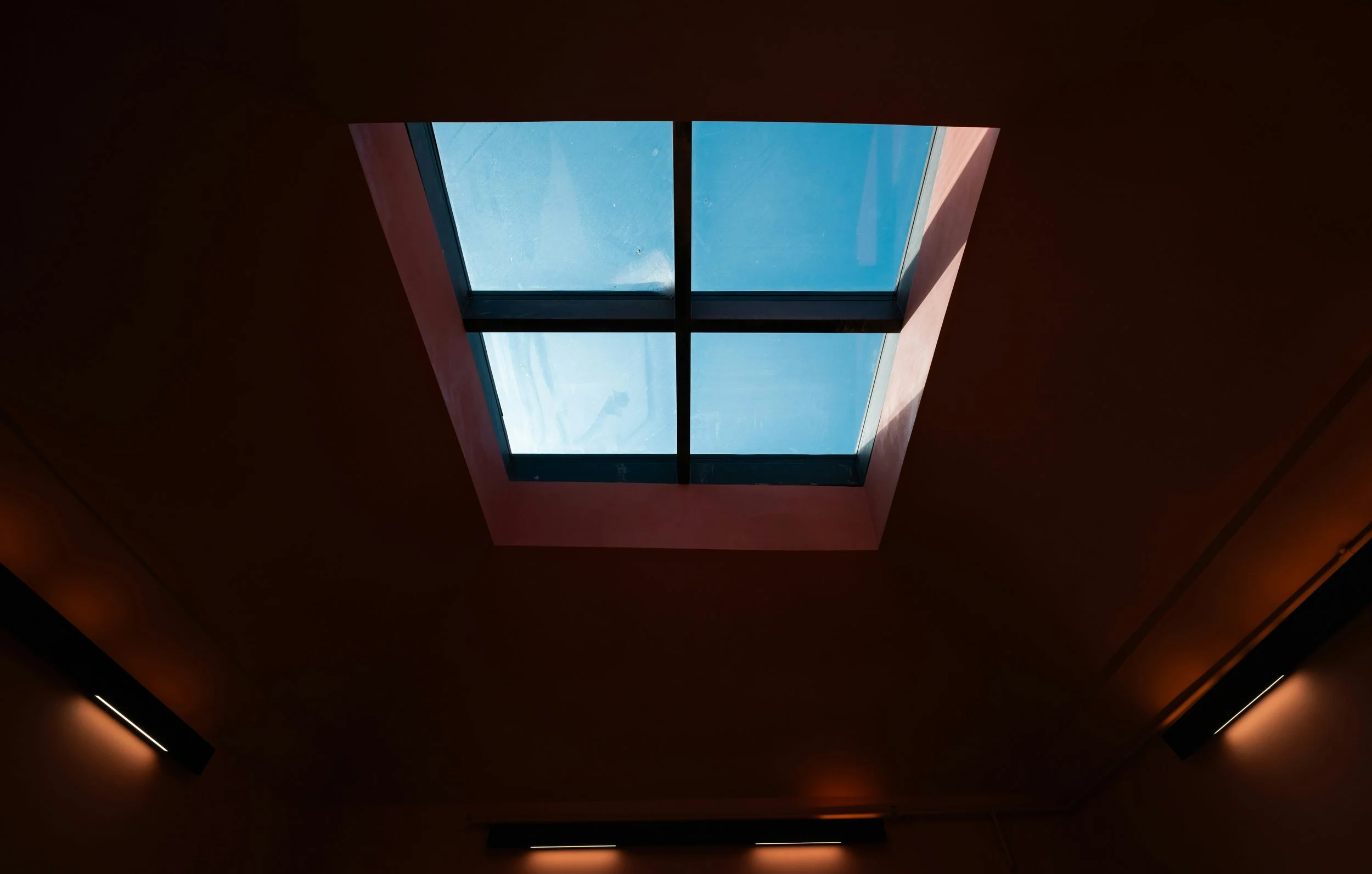 Interior ceiling view with large skylight window showing blue sky and clouds, surrounded by dark brown walls with wall-mounted lights.