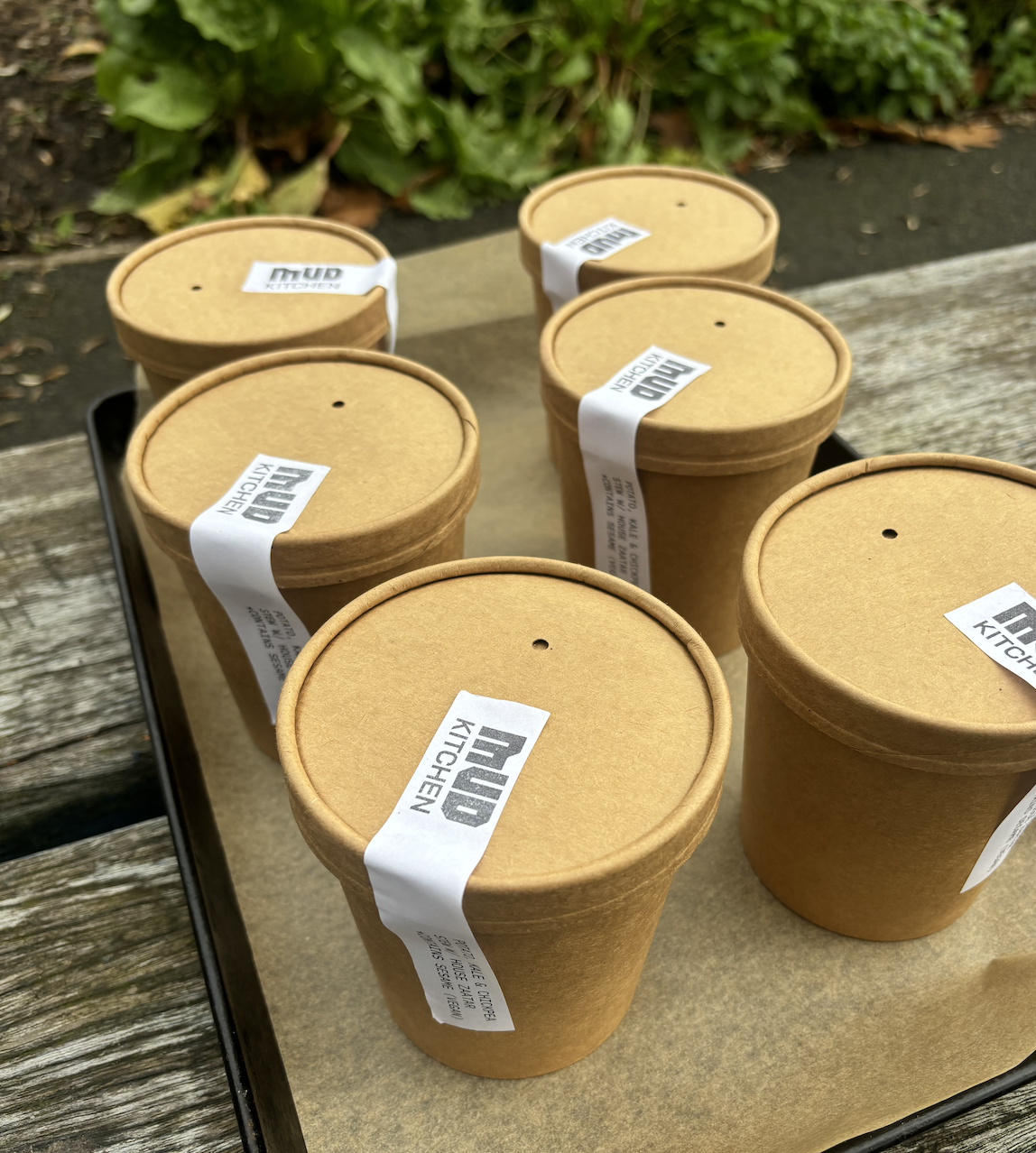 Six brown paper cups with lids on a black tray outside, with green plants and wooden bench in the background.