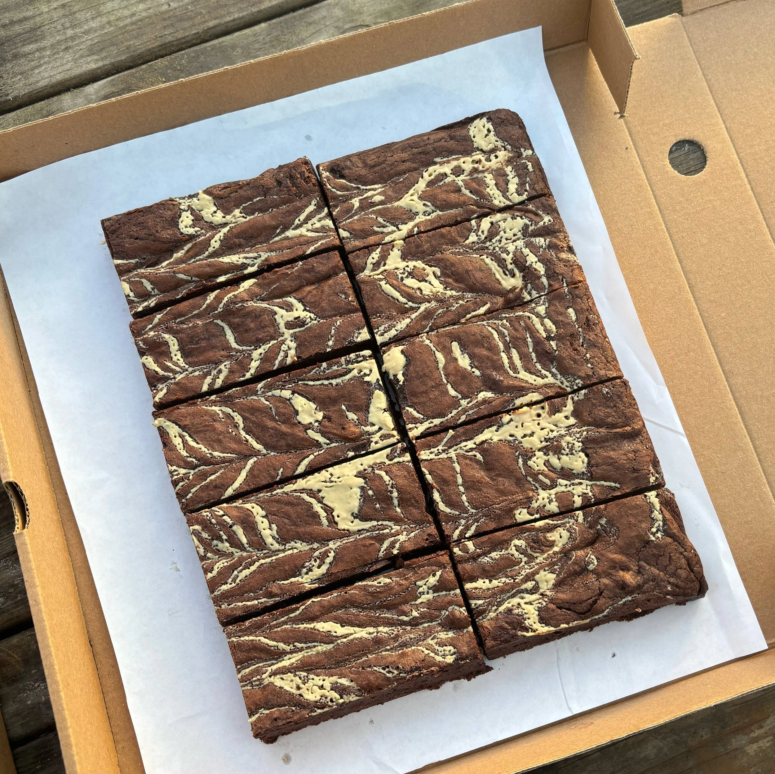 A box of large, rectangular chocolate brownies with white icing drizzle, arranged in three rows of four pieces on parchment paper.