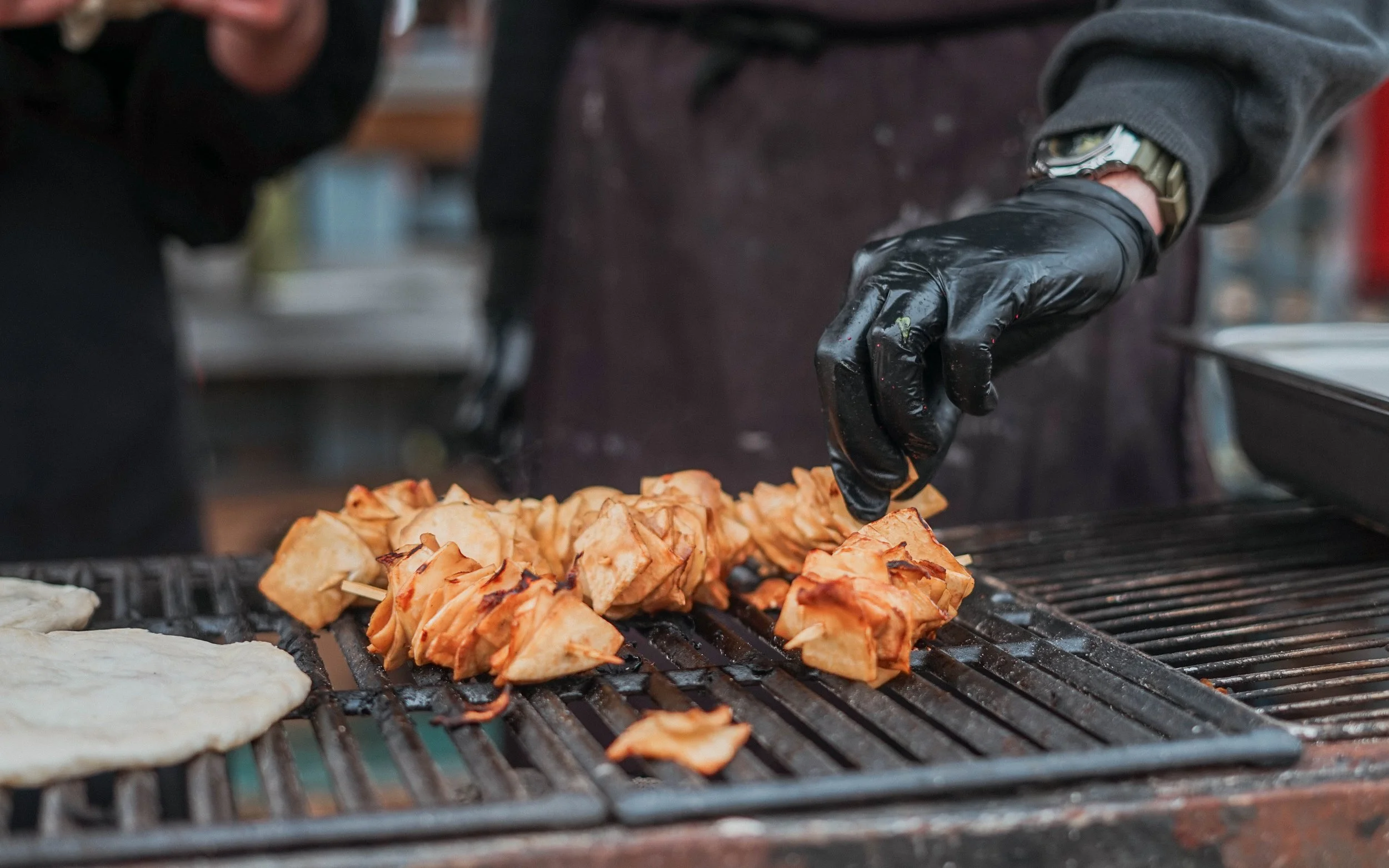 Person wearing black gloves cooking vegan skewers on a grill, with a flatbread nearby.