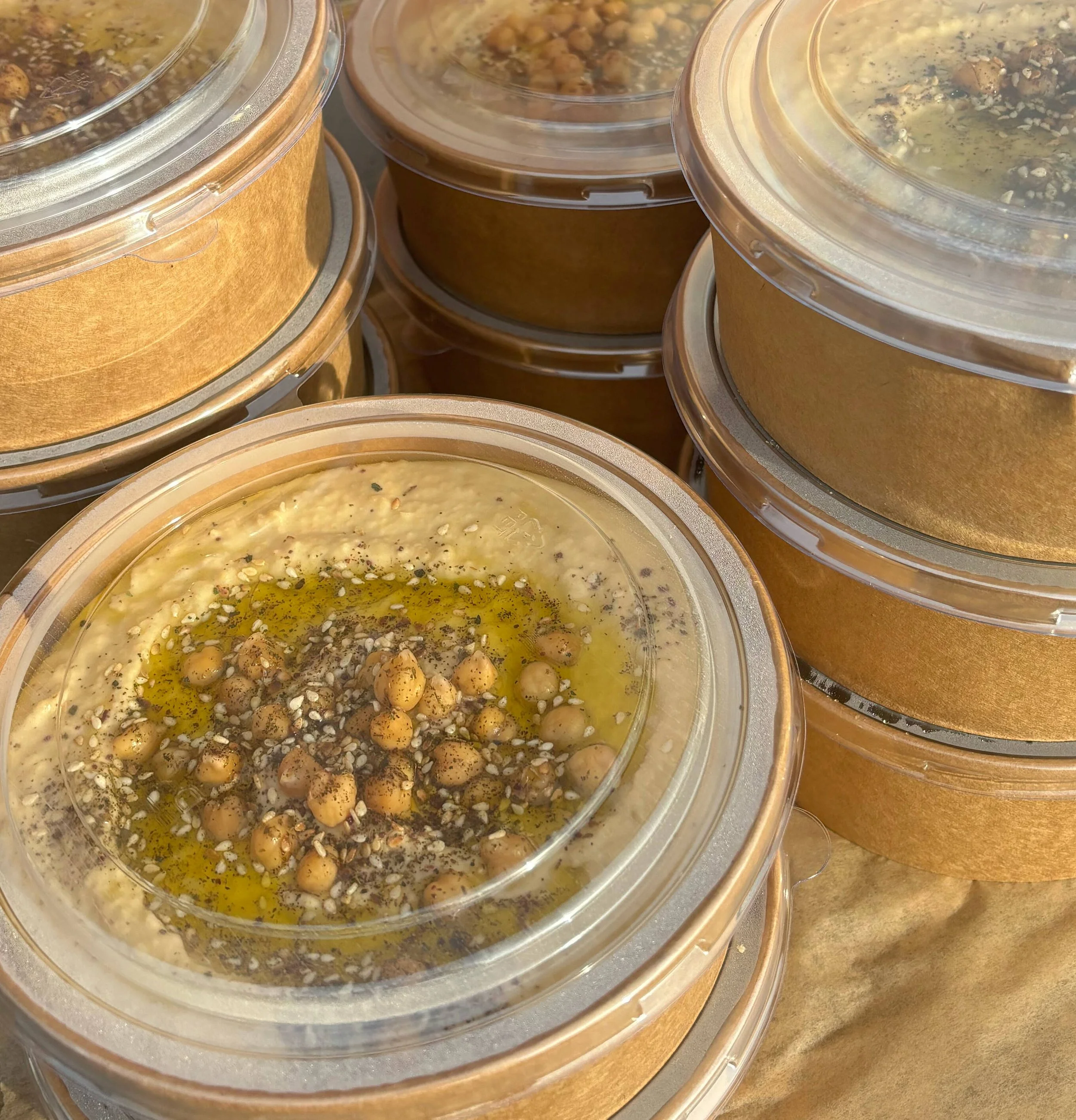 Plastic containers with hummus topped with chickpeas, sesame seeds, and olive oil, stacked on a surface.