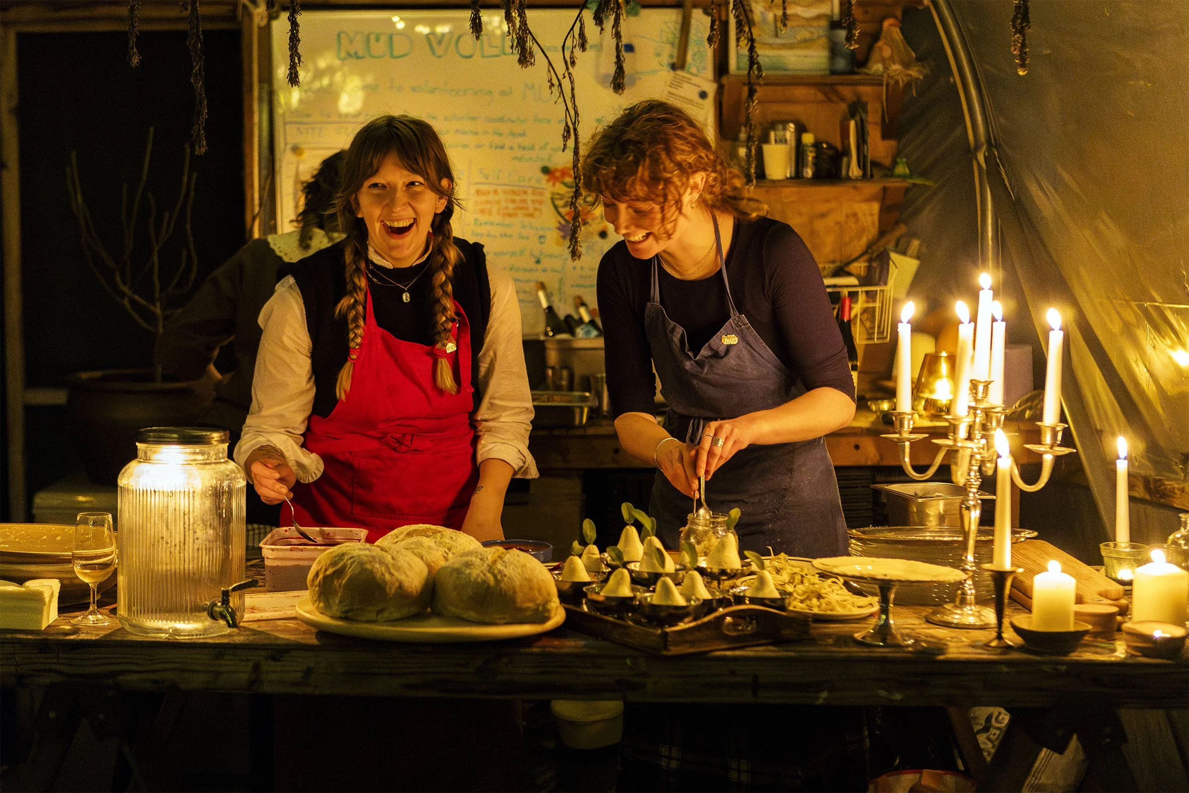 Two women in a warmly lit, cozy kitchen or dining area, preparing food and enjoying each other's company amid candles and decorative elements.