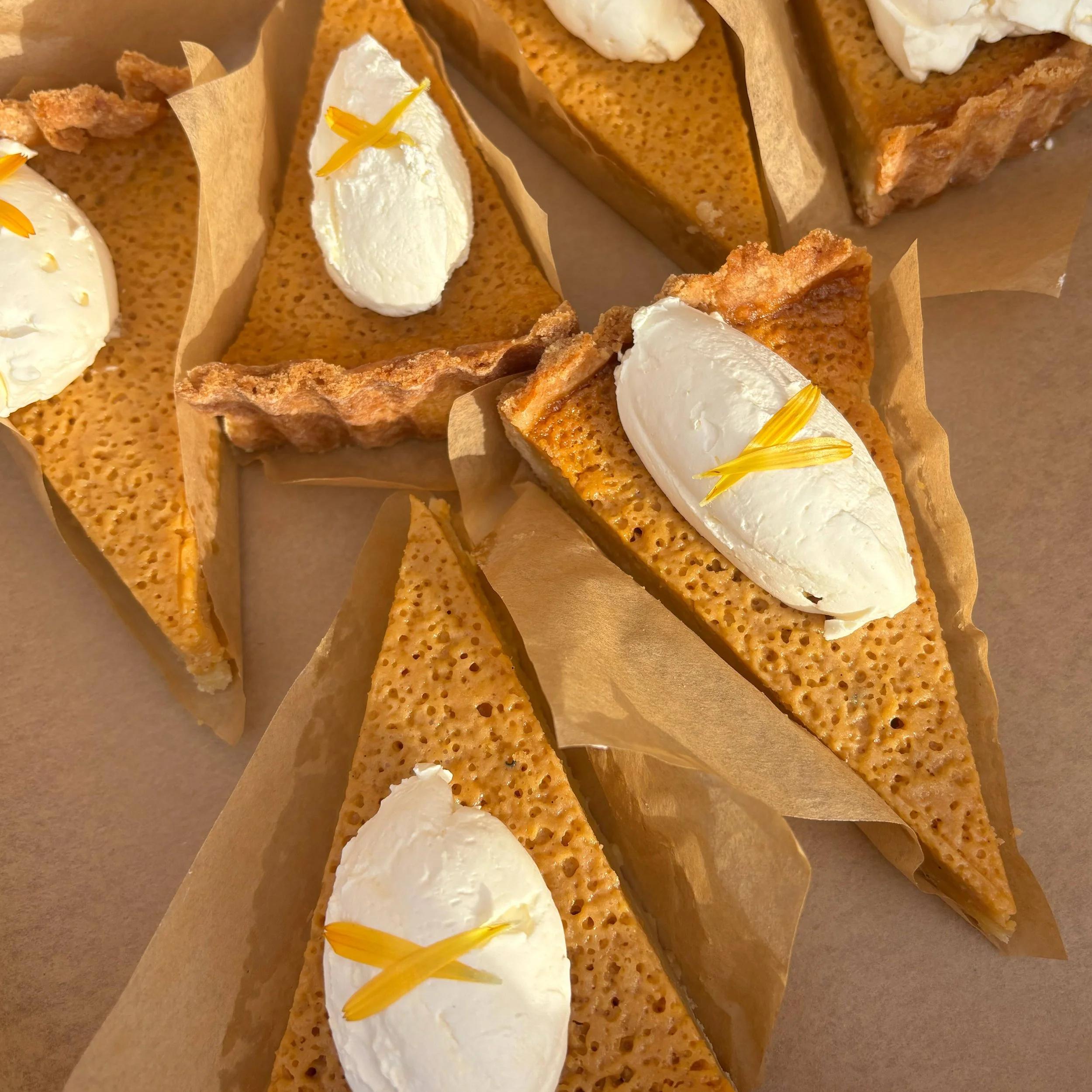 Slices of caramelized honeycomb toffee with dollops of whipped cream and yellow edible flower petals on top, arranged on parchment paper.