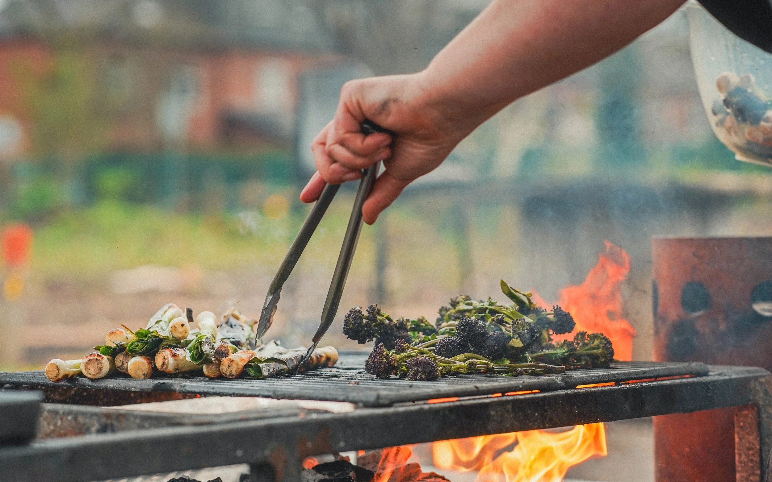 Hand using tongs to cook vegetables on a barbecue grill with flames visible underneath.