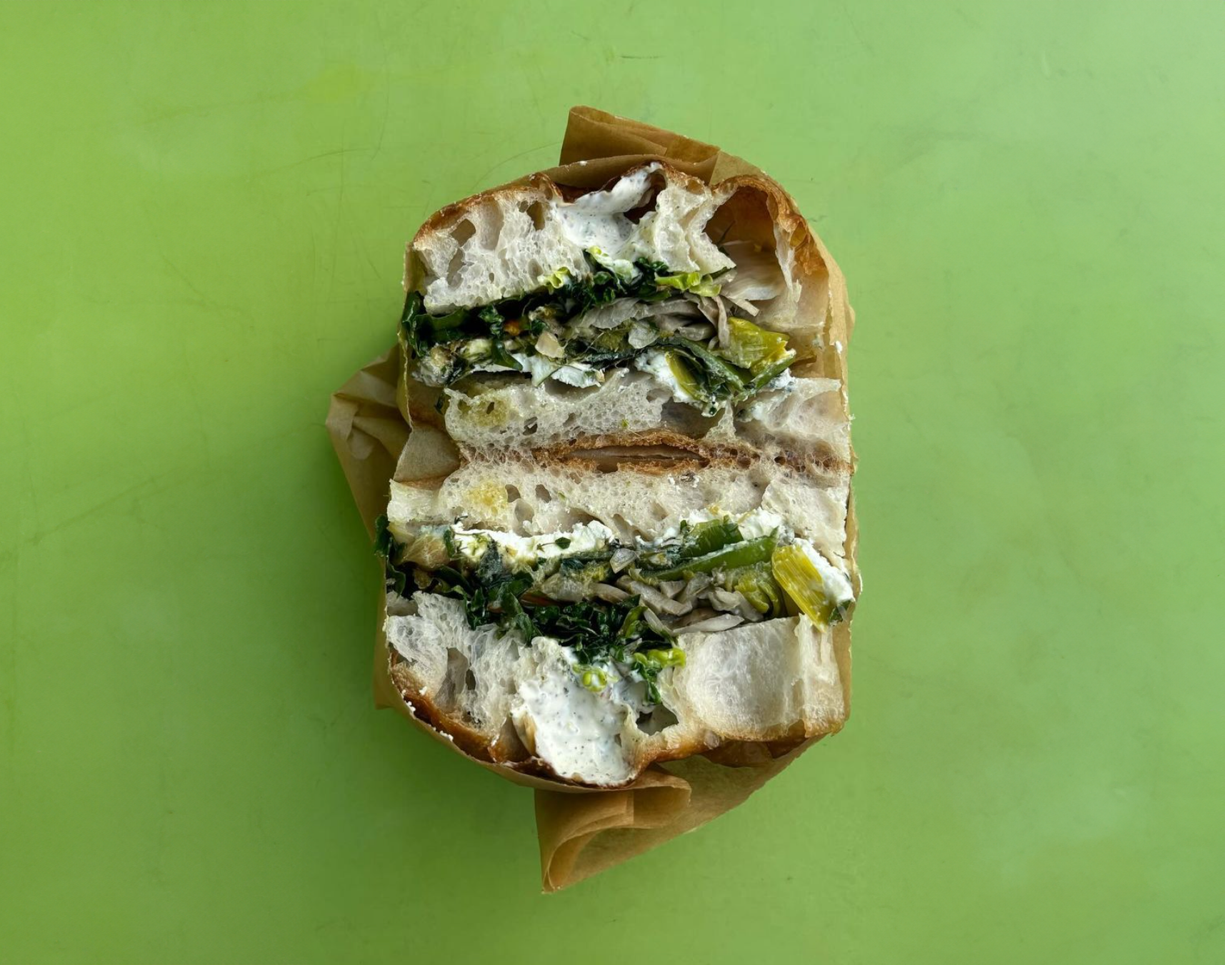 Cross-section of a sandwich with leafy greens, cheese, and creamy sauce in a bread roll on a green background.