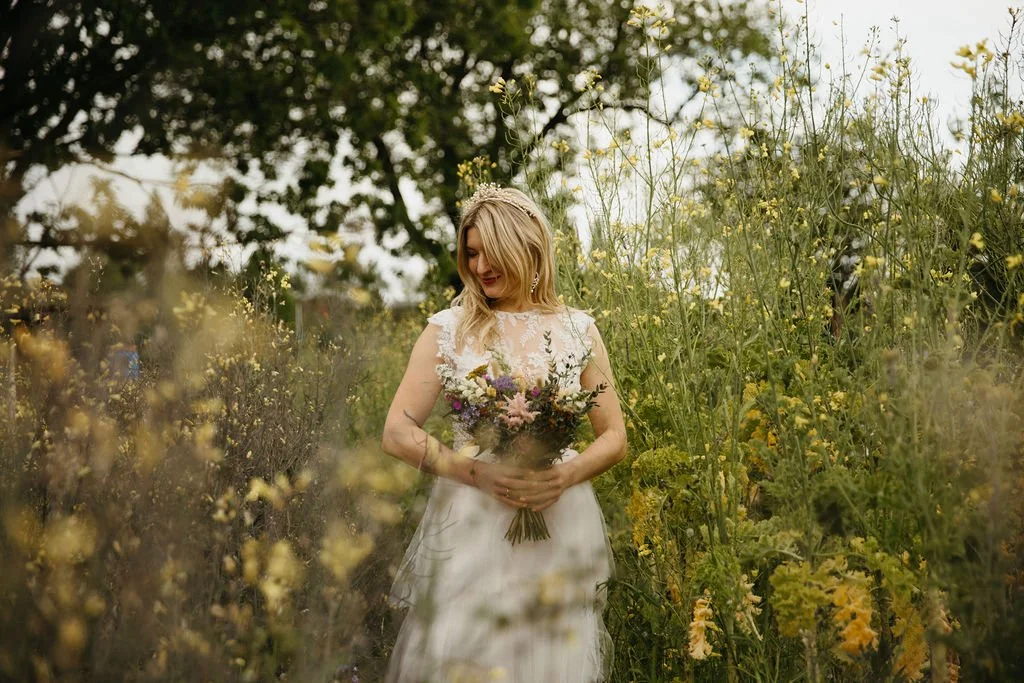 A woman in a white wedding dress standing in a field of yellow and green wildflowers, holding a bouquet of mixed flowers, with trees in the background.