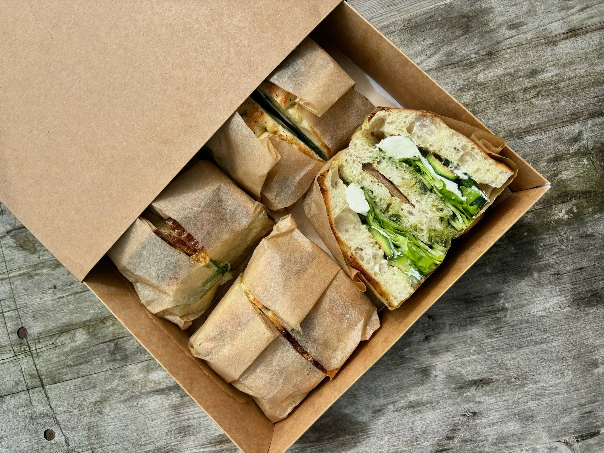 Open cardboard box containing multiple wrapped sandwiches and one unwrapped sandwich with lettuce, cucumber, and slices of chicken on a wooden surface.