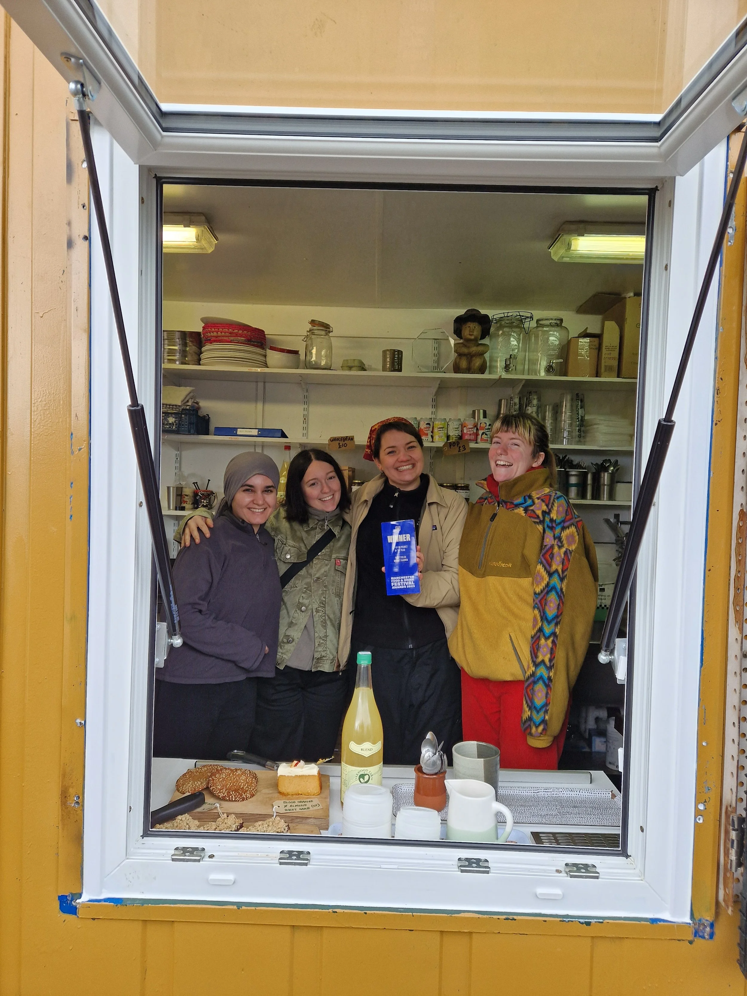 Four women smiling and standing together inside a kitchen, seen through a large window. They are holding an award.