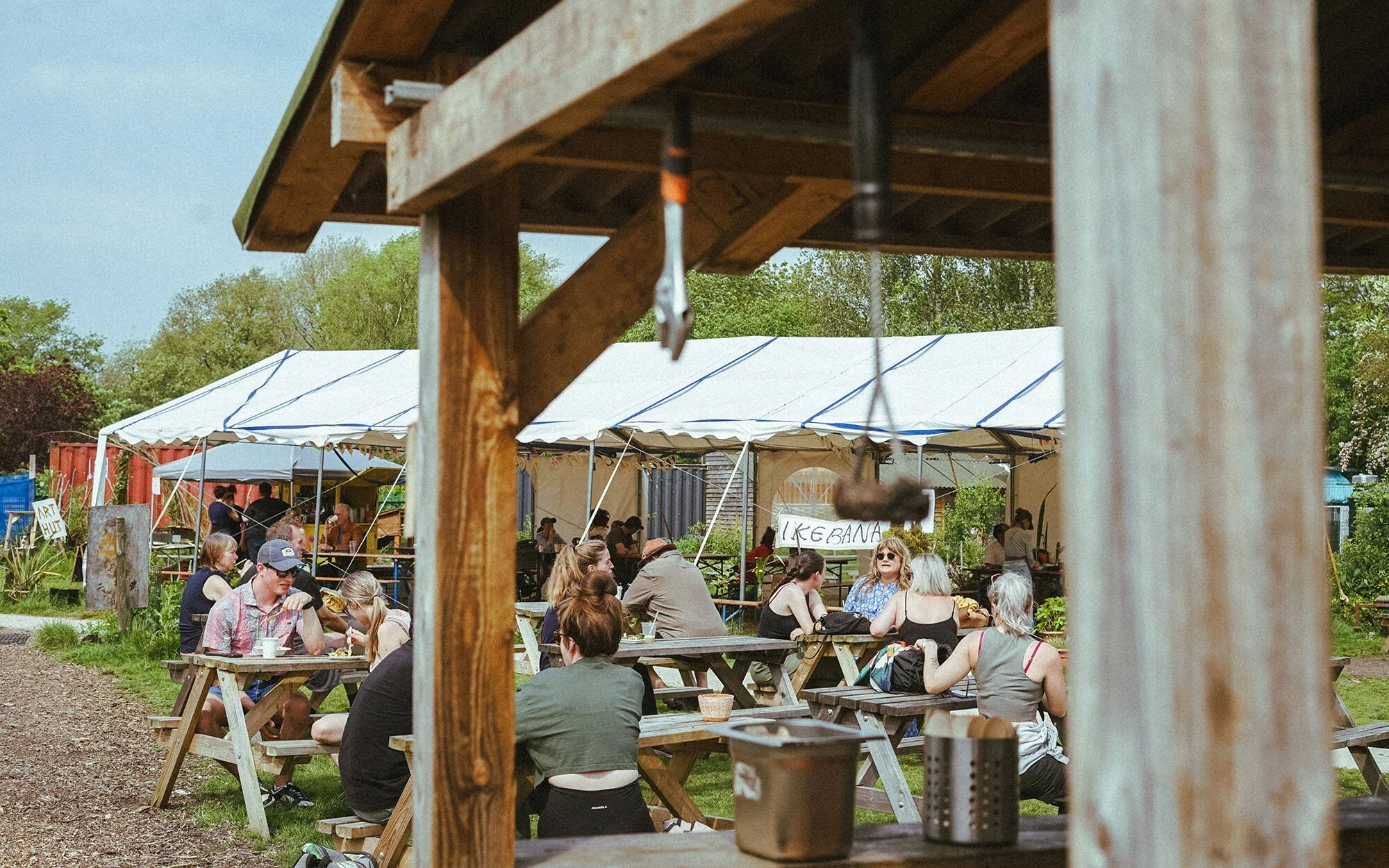 Outdoor gathering with people sitting at picnic tables under a large white canopy, surrounded by greenery and wooden structures.