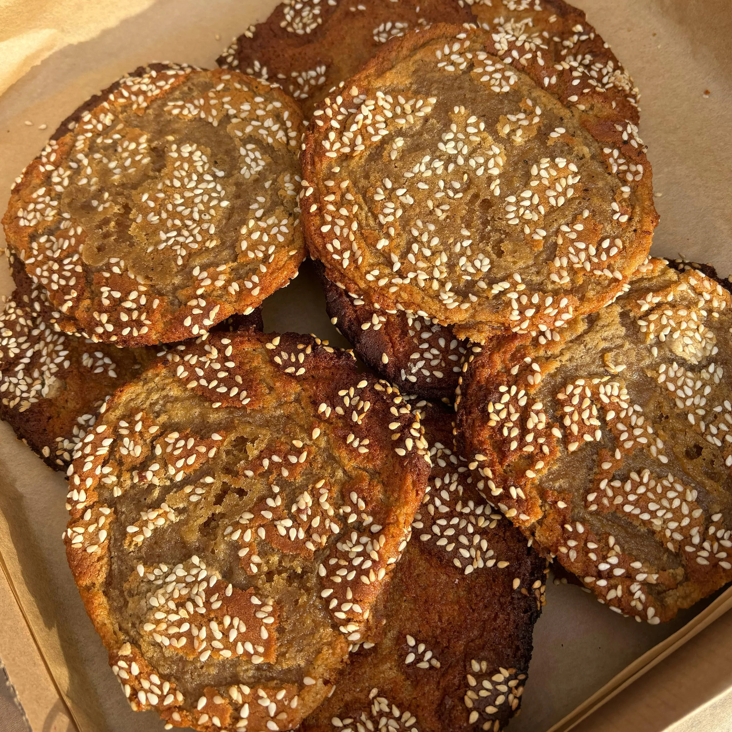 Baked cookies with a golden-brown crust, topped with white sesame seeds, in a paper-lined tray.