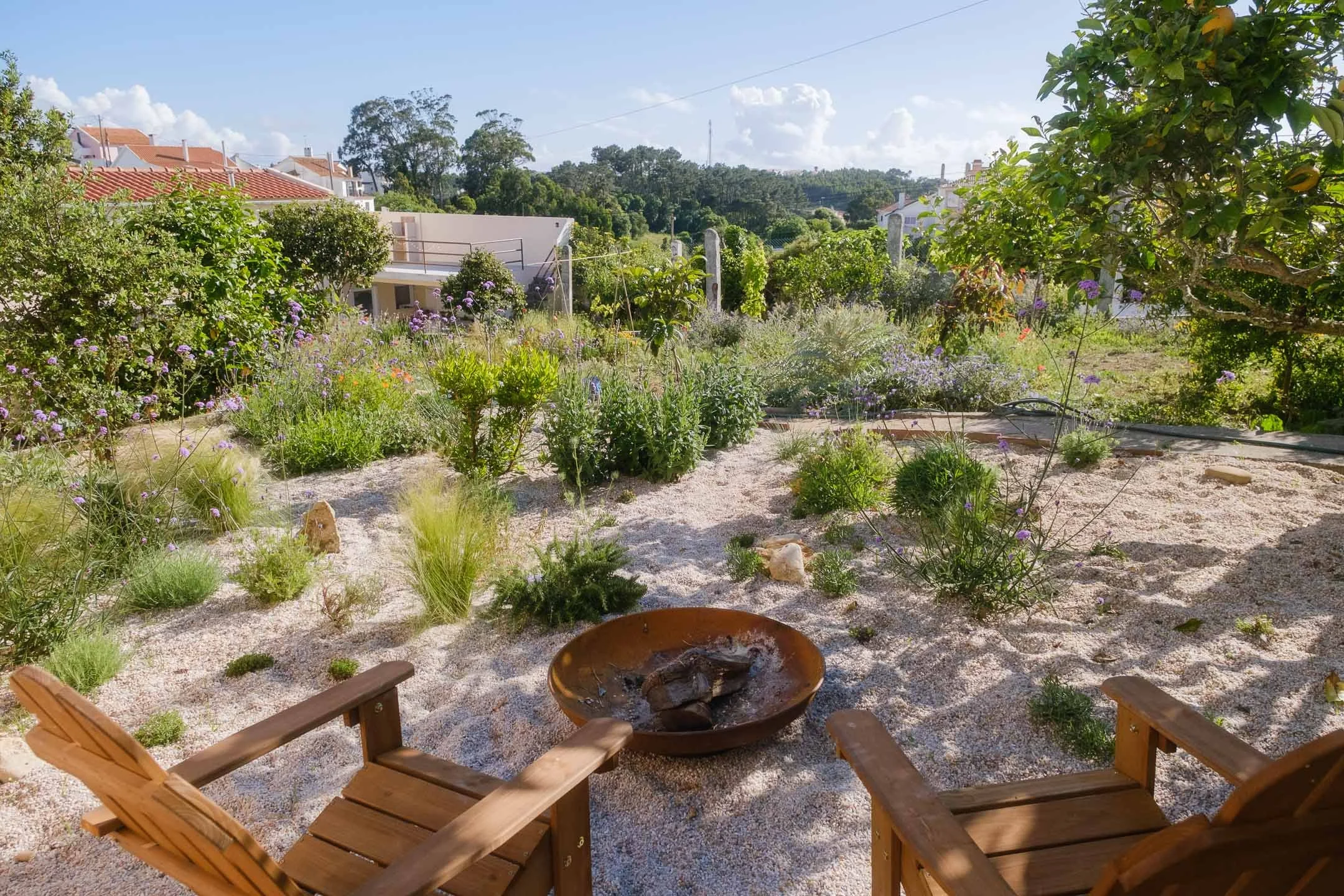 Two wooden chairs facing a fire pit in a natural gravel garden with native portuguese plants, flowers, and trees under a blue sky with clouds.