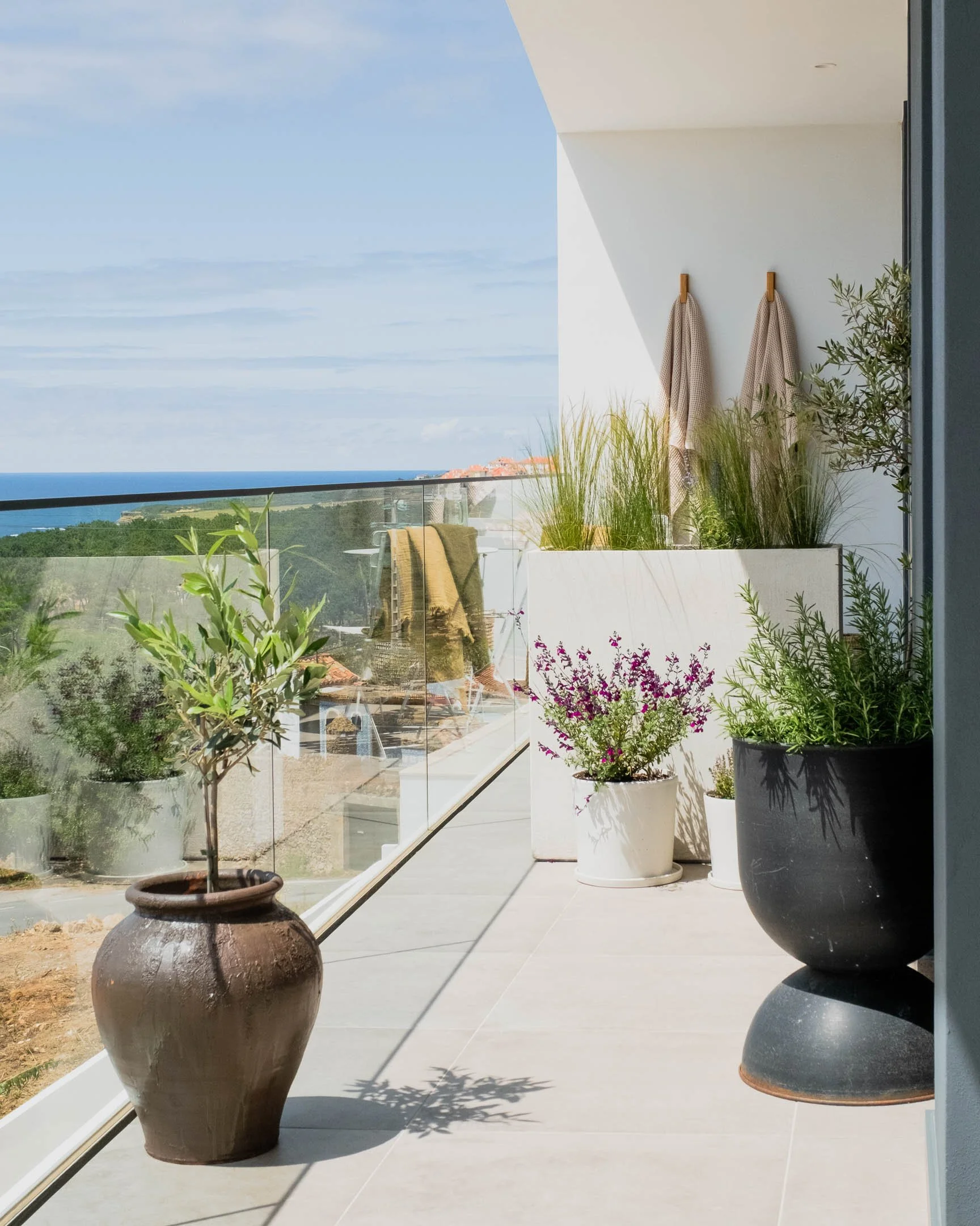 Balcony with potted plants and towels, overlooking ocean and greenery in the distance, sunny day.