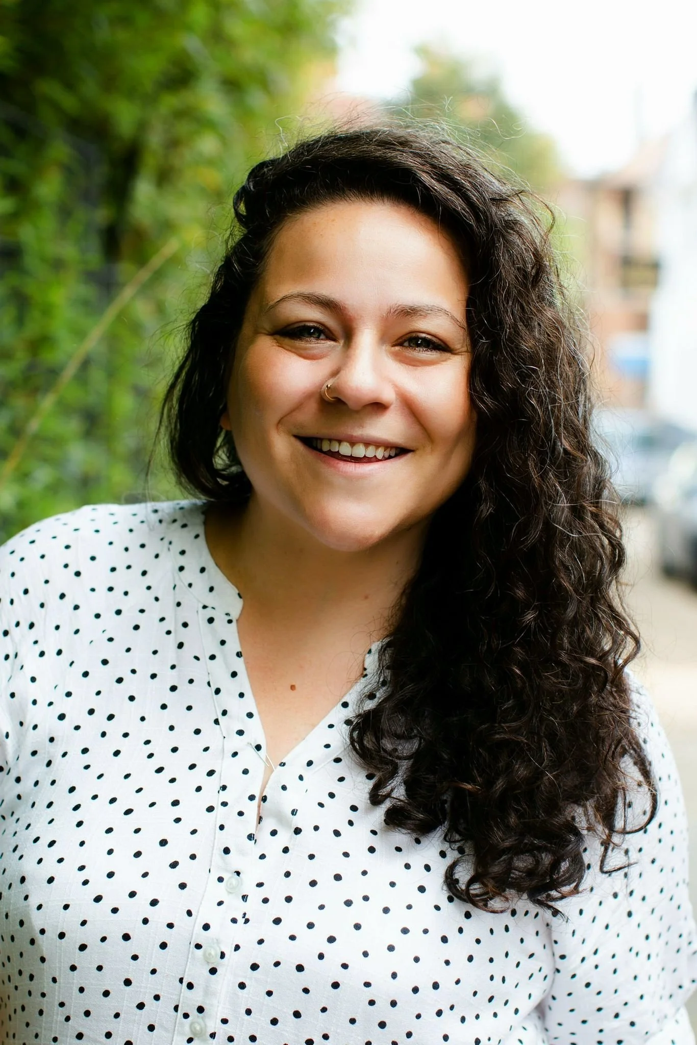 A woman with dark curly hair, smiling, wearing a white shirt with black polka dots, standing outdoors with greenery and blurred buildings in the background.