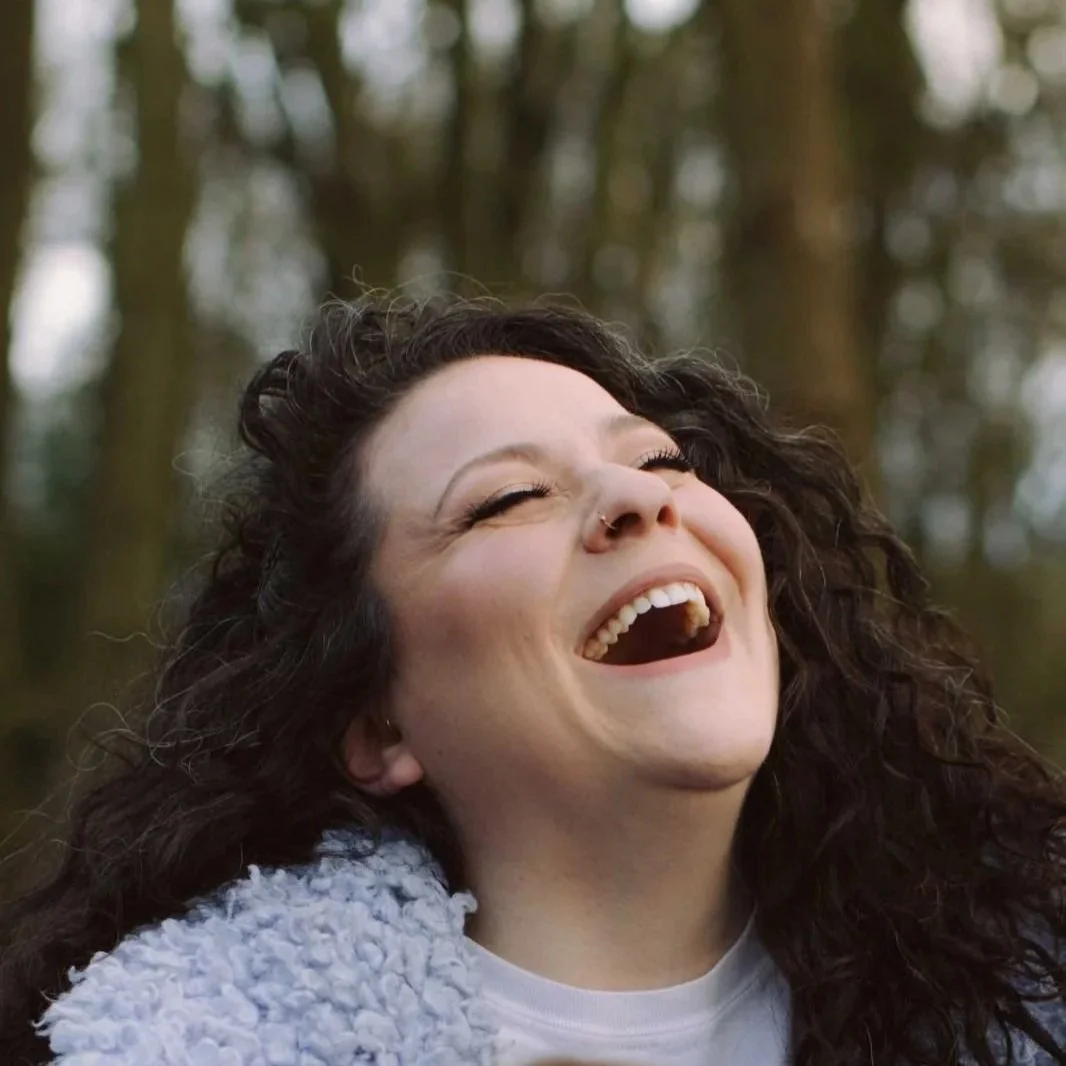 A person with curly dark hair smiling and laughing outdoors in a wooded area.