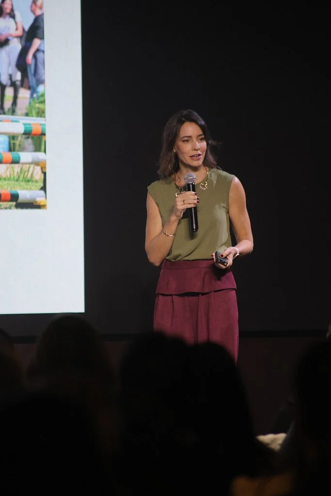 Mulher falando em um microfone durante uma apresentação, com um slide ao fundo.