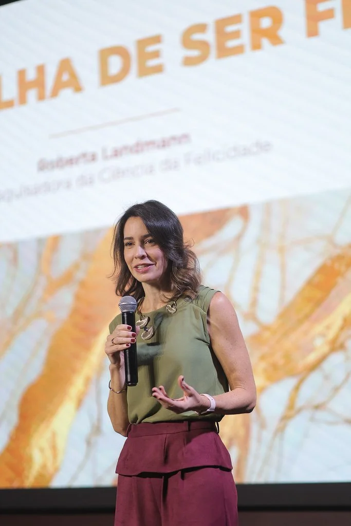 Mulher falando em um palco com microfone, fundo com texto em português e cores quentes.