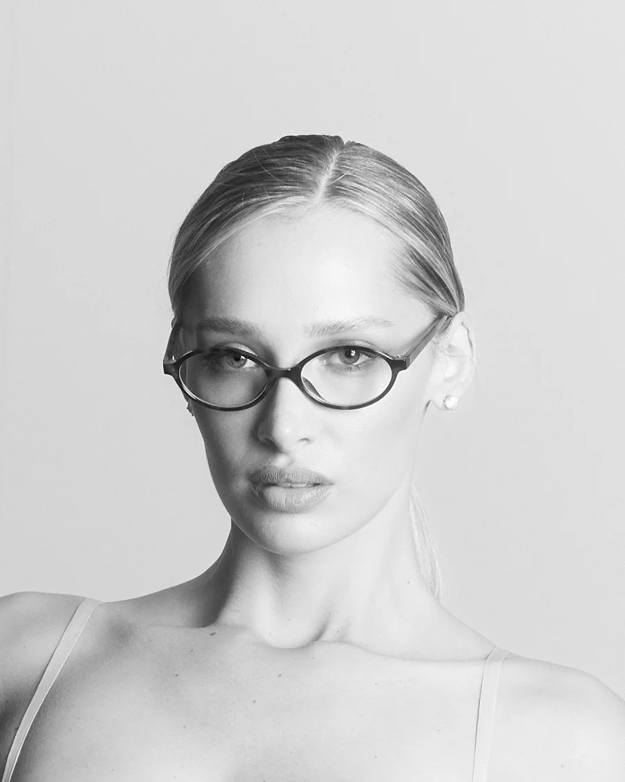 Black and white portrait of a woman with glasses, light-colored hair pulled back, and wearing a spaghetti strap top, against a plain background.