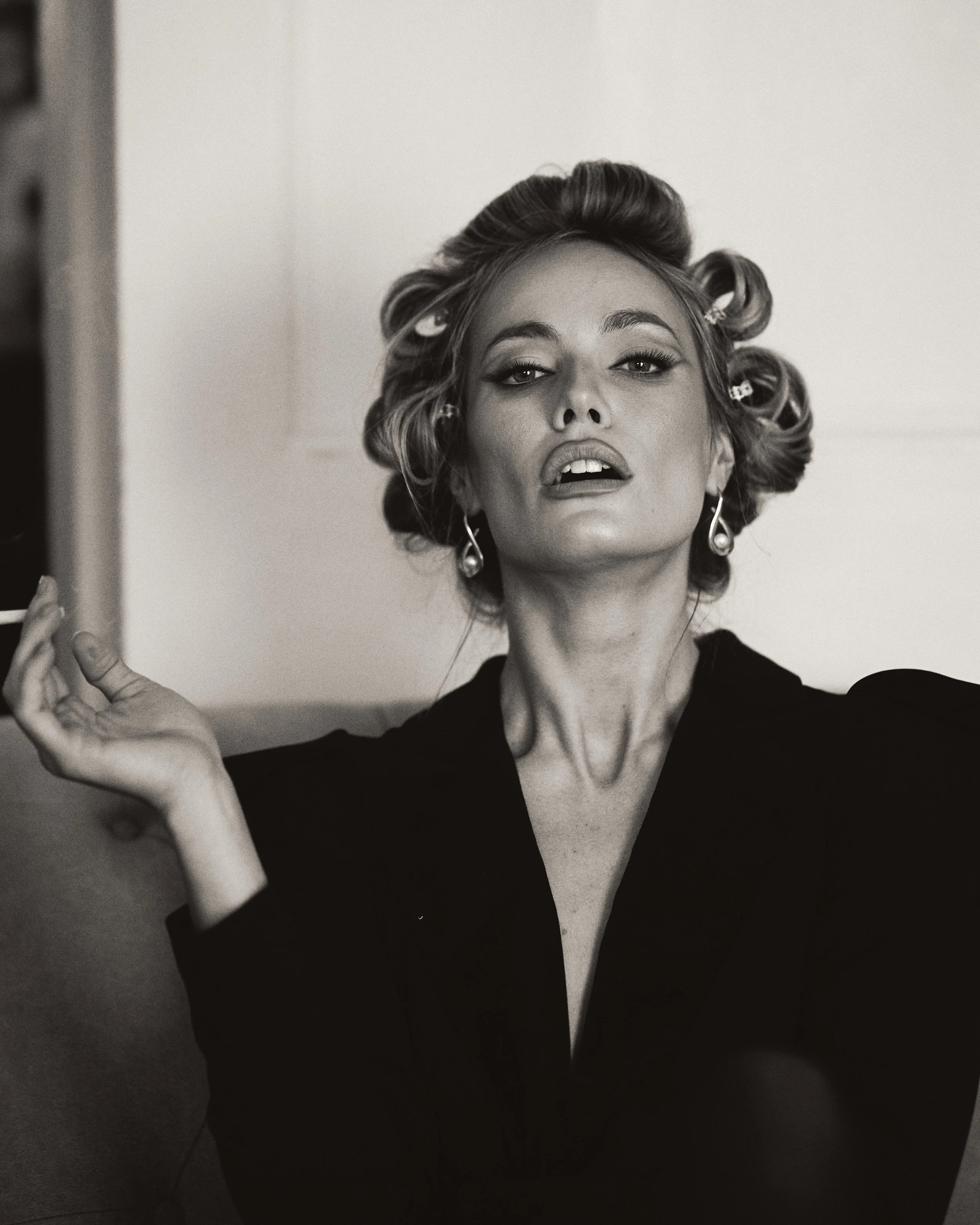 Black-and-white photo of a woman with vintage hair rollers, wearing earrings and a dark top, looking confidently at the camera.