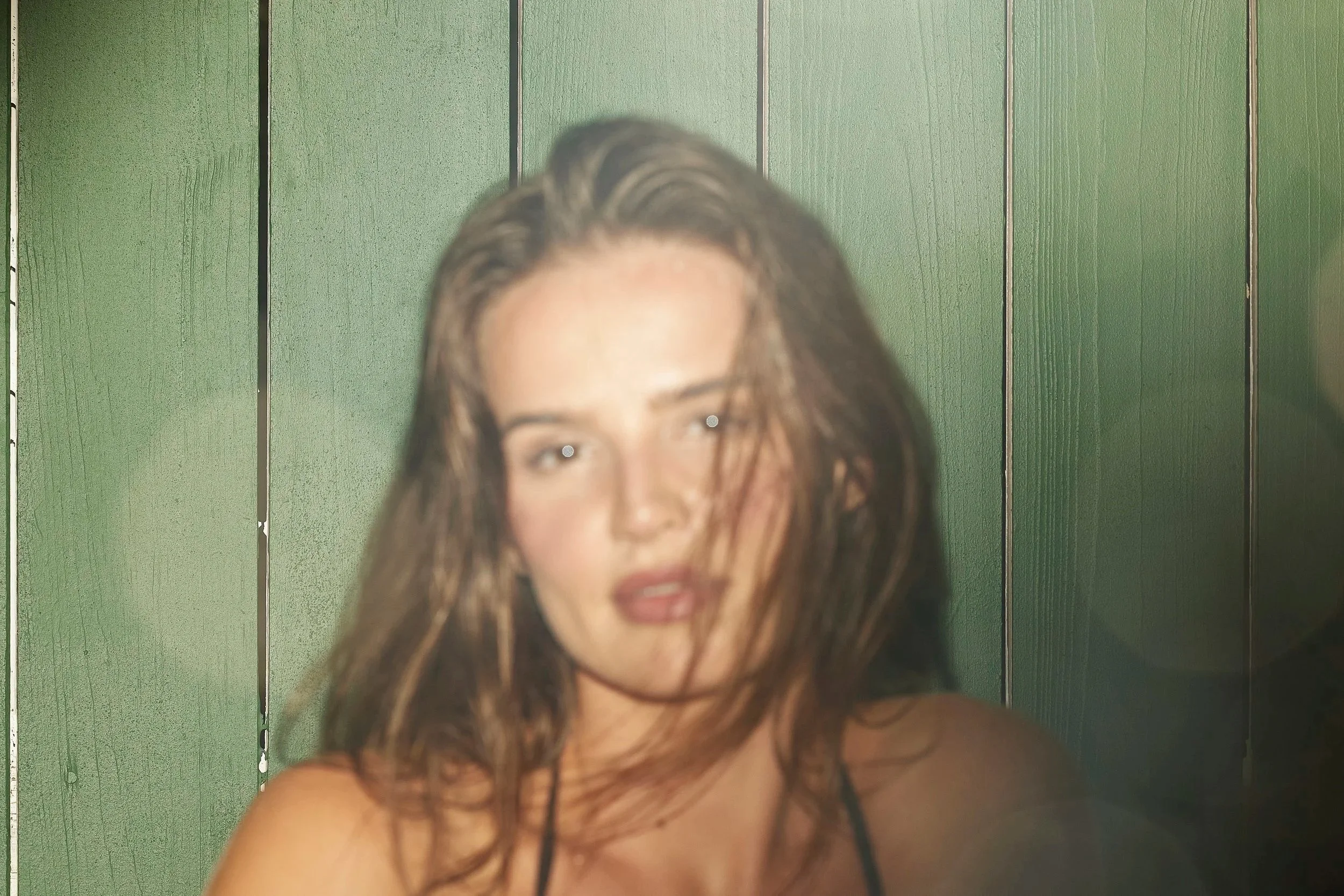 A woman with long brown hair, slightly tousled, standing in front of a green wooden paneled wall, wearing a black spaghetti strap top.