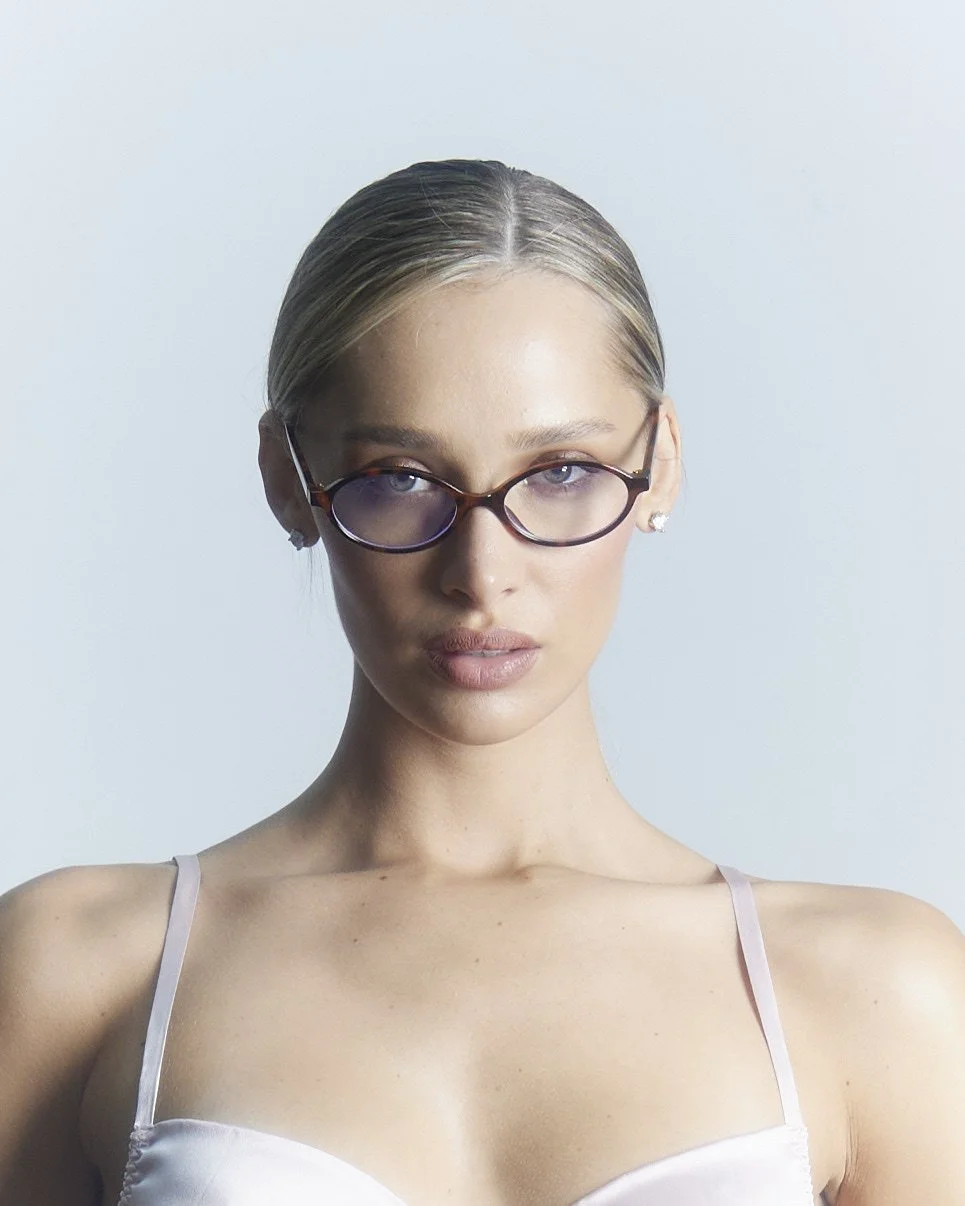 A woman with blonde hair slicked back, wearing round glasses and a white spaghetti strap top, looking directly at the camera against a plain light background.