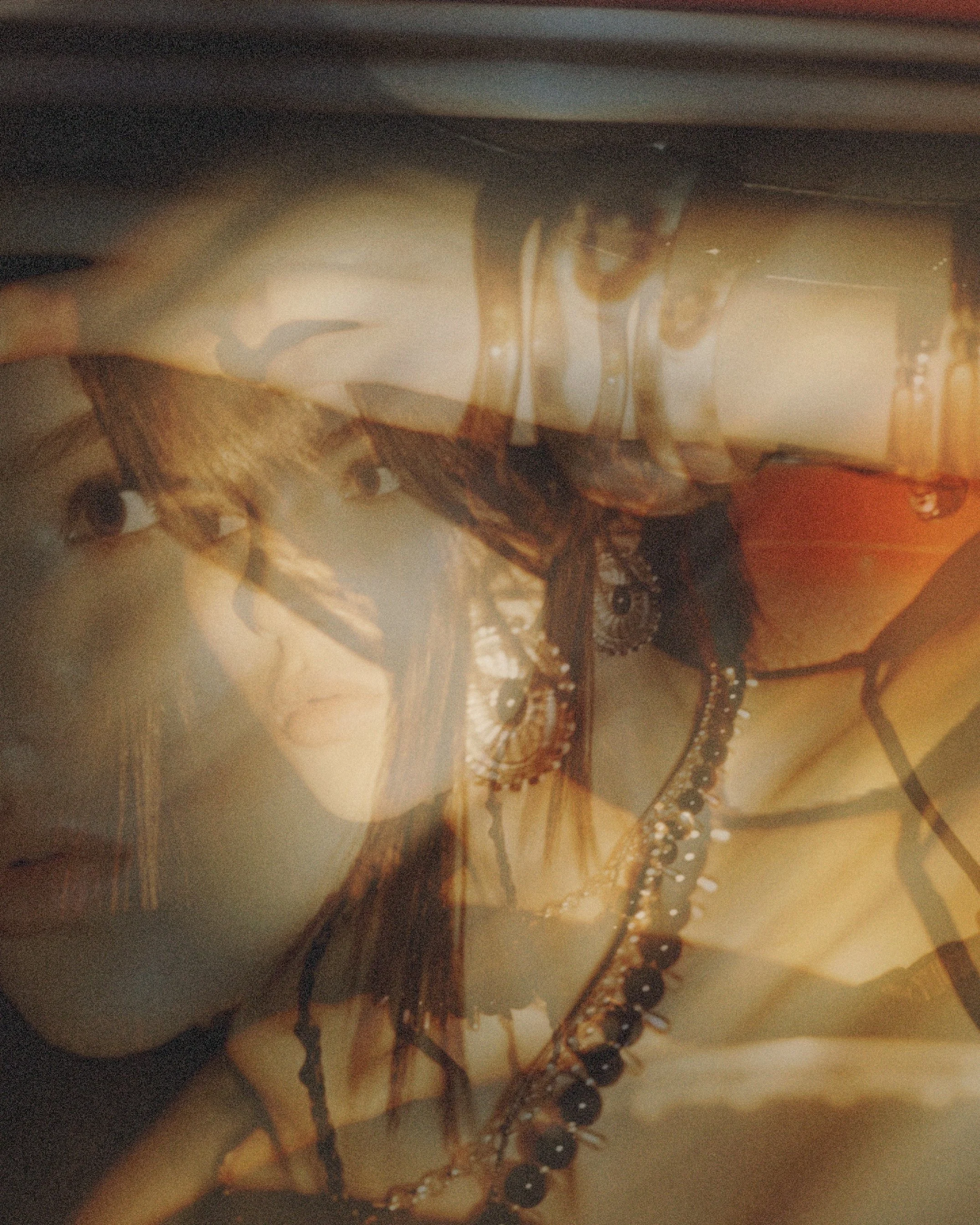 A double exposure photograph of a woman's face and a woman with long hair, wearing jewelry, and holding her glasses. The images are layered, creating an artistic and abstract effect.