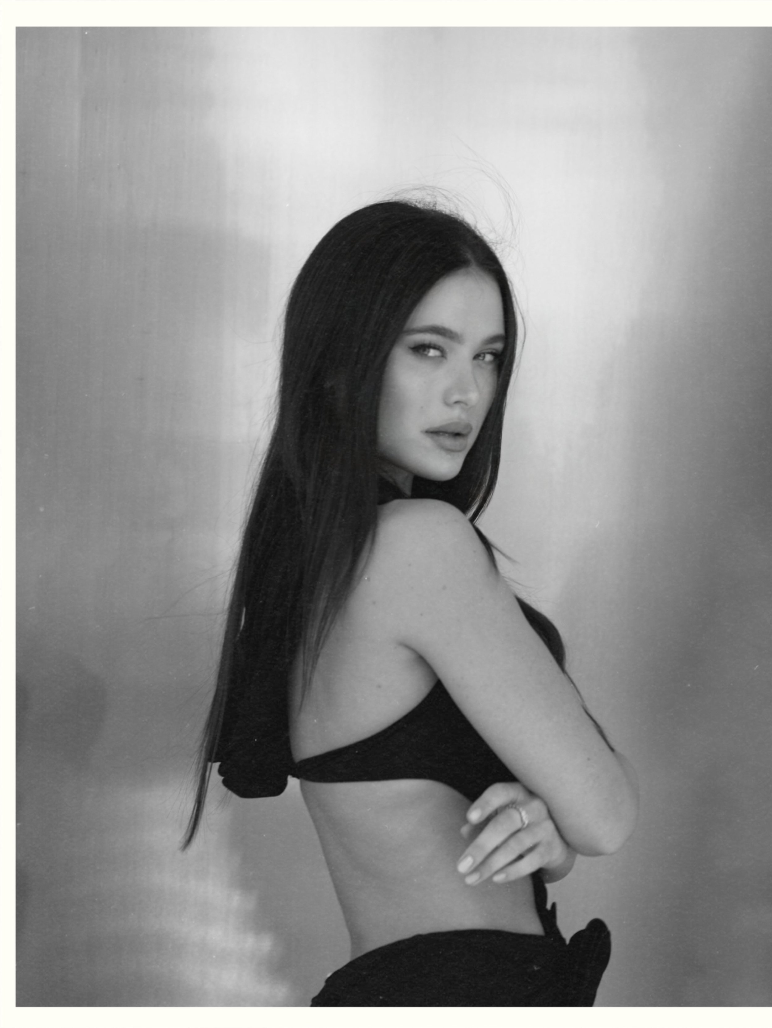 Black and white photo of a woman with long dark hair, wearing a revealing top, looking over her shoulder with an expression that is both confident and sultry.