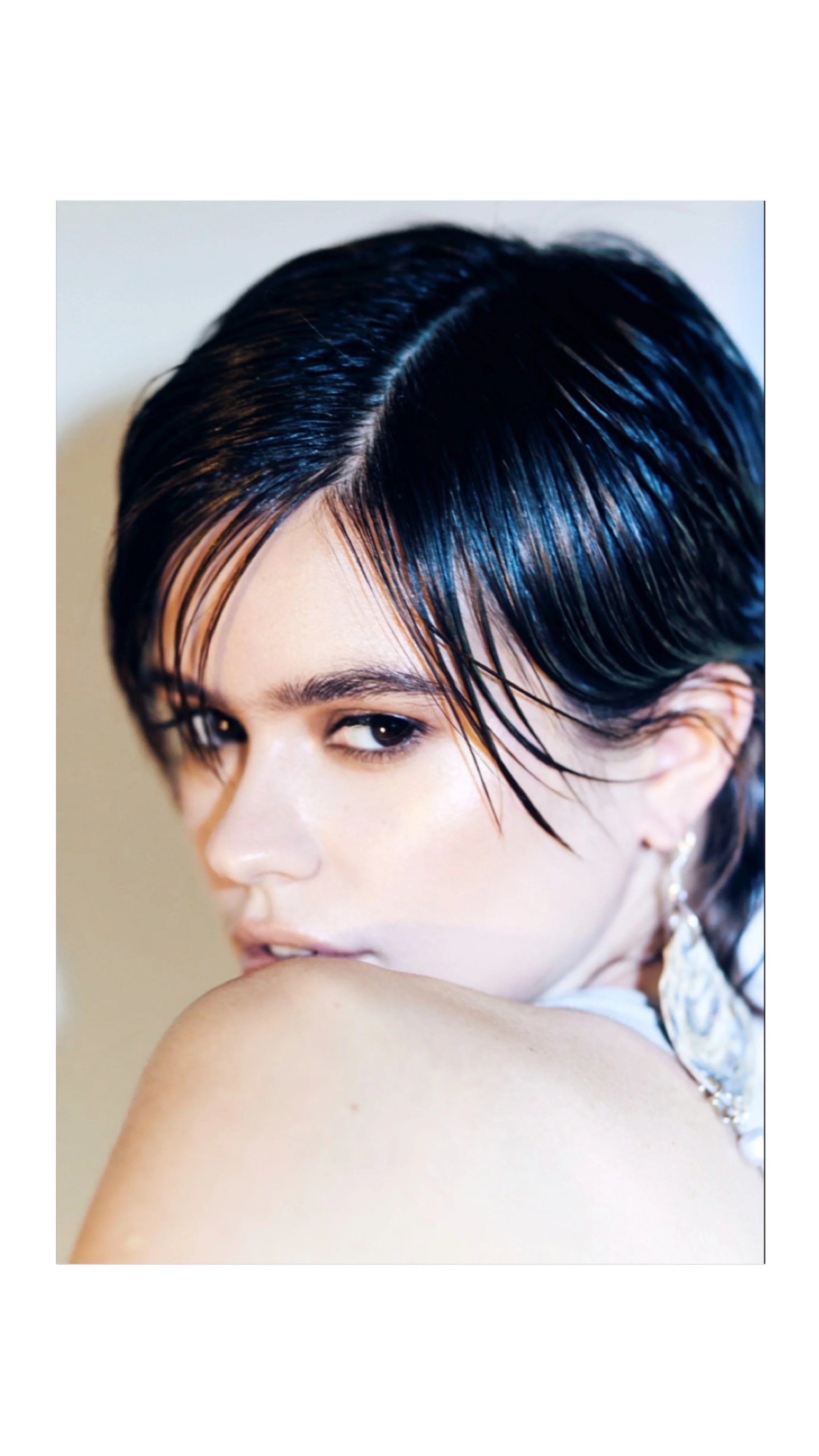 Close-up portrait of a young woman with wet, dark hair styled in a sleek bob, looking over her shoulder with a neutral expression.