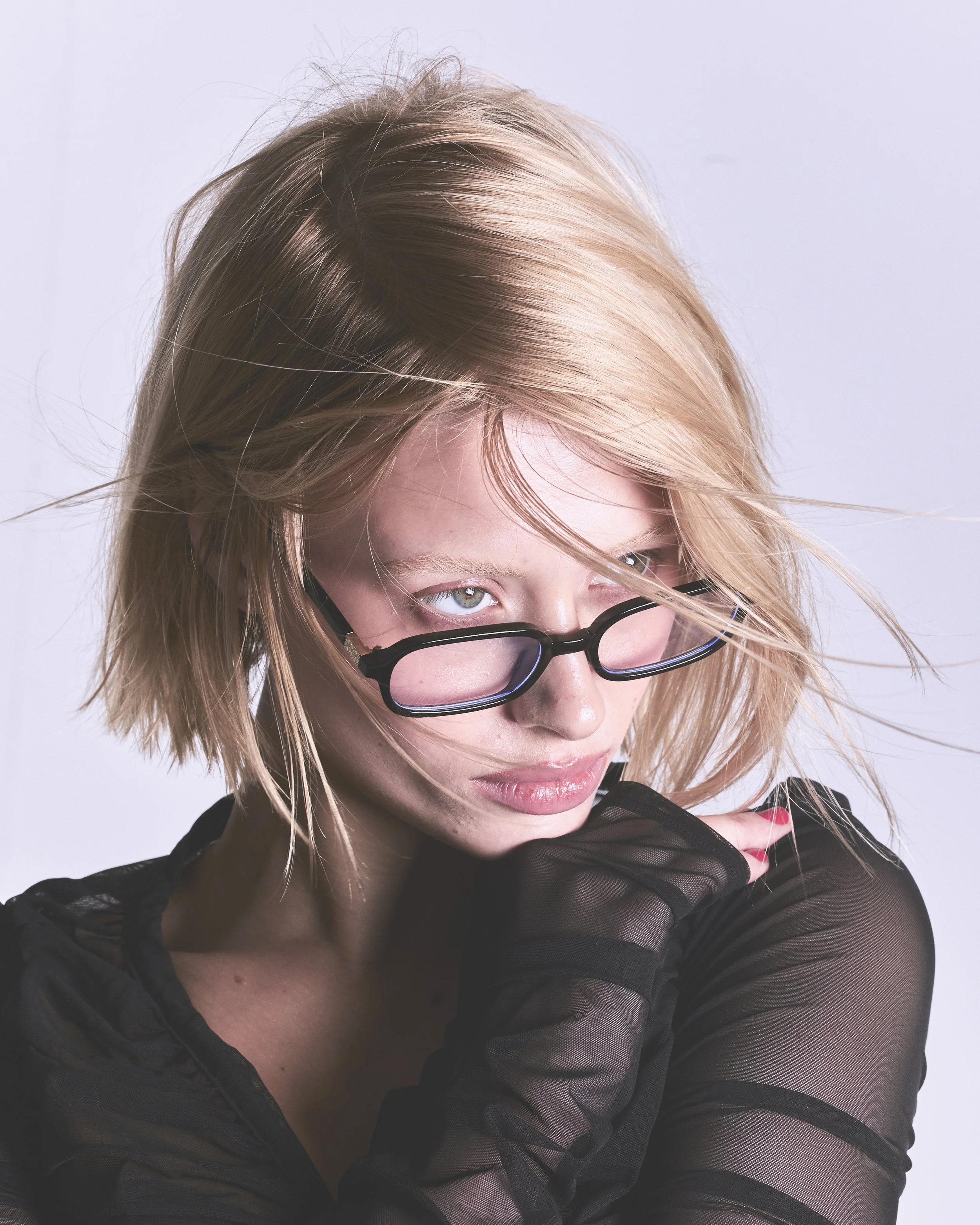 A woman with blonde hair, wearing black-rimmed glasses, and a black sheer top. She has her hand raised to her face and is looking slightly downward.