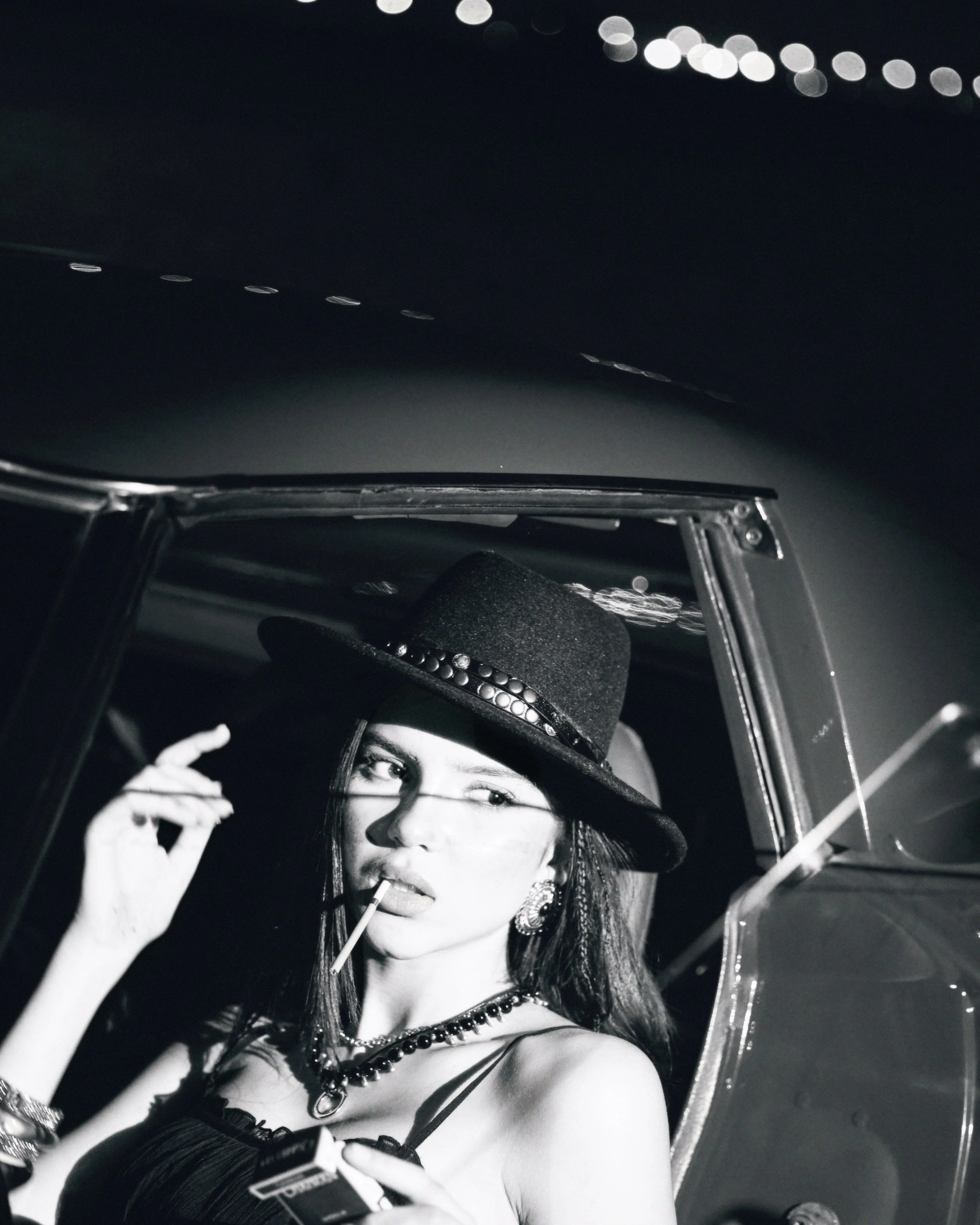 A woman sitting in a car, wearing a fedora hat and jewelry, holding a cigarette in her mouth and a pack of cigarettes in her hand, with a city street and lights in the background.