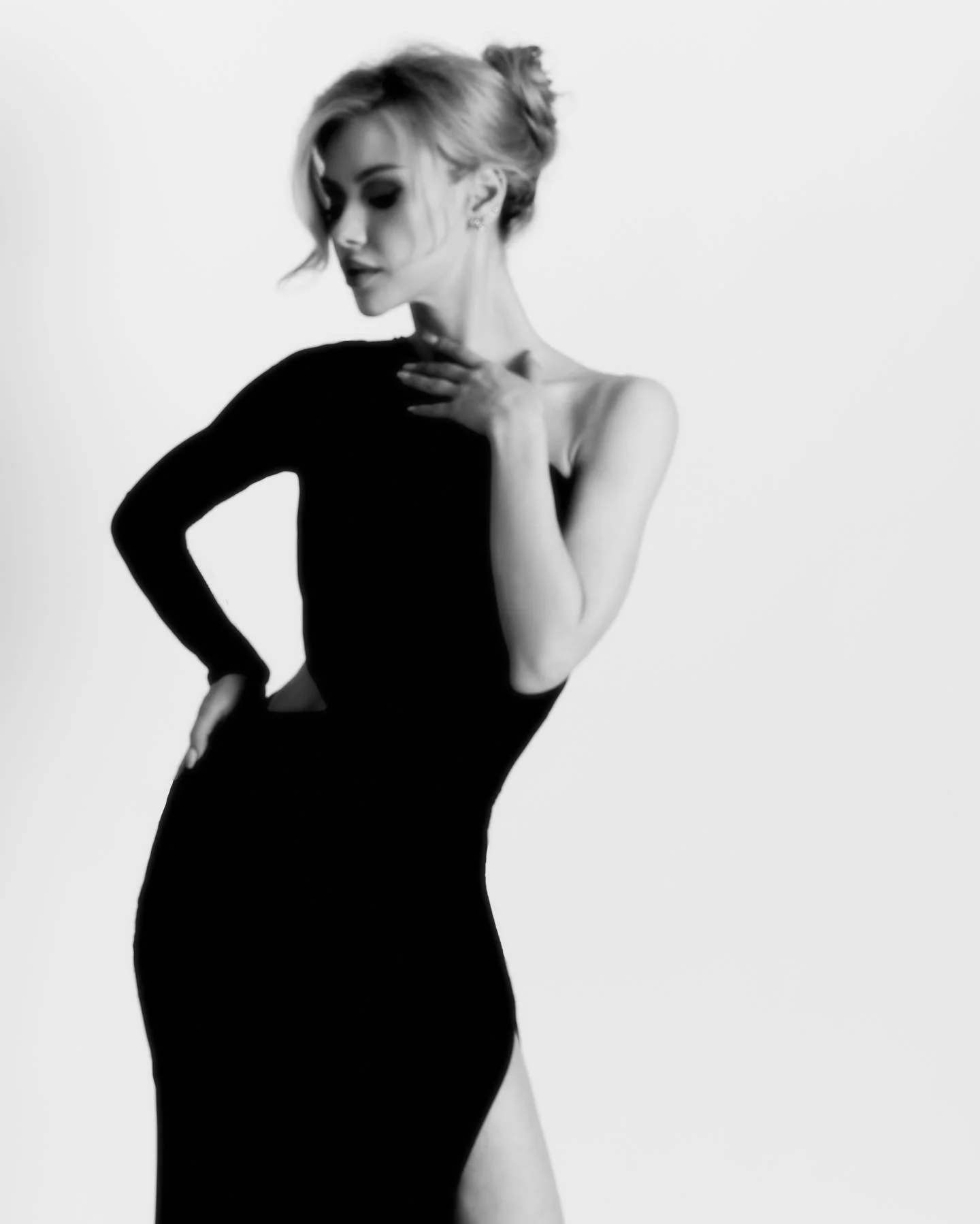 A woman in a black dress with one shoulder exposed, posing with her hand on her chest and her head slightly bowed, photographed in black and white against a plain background.