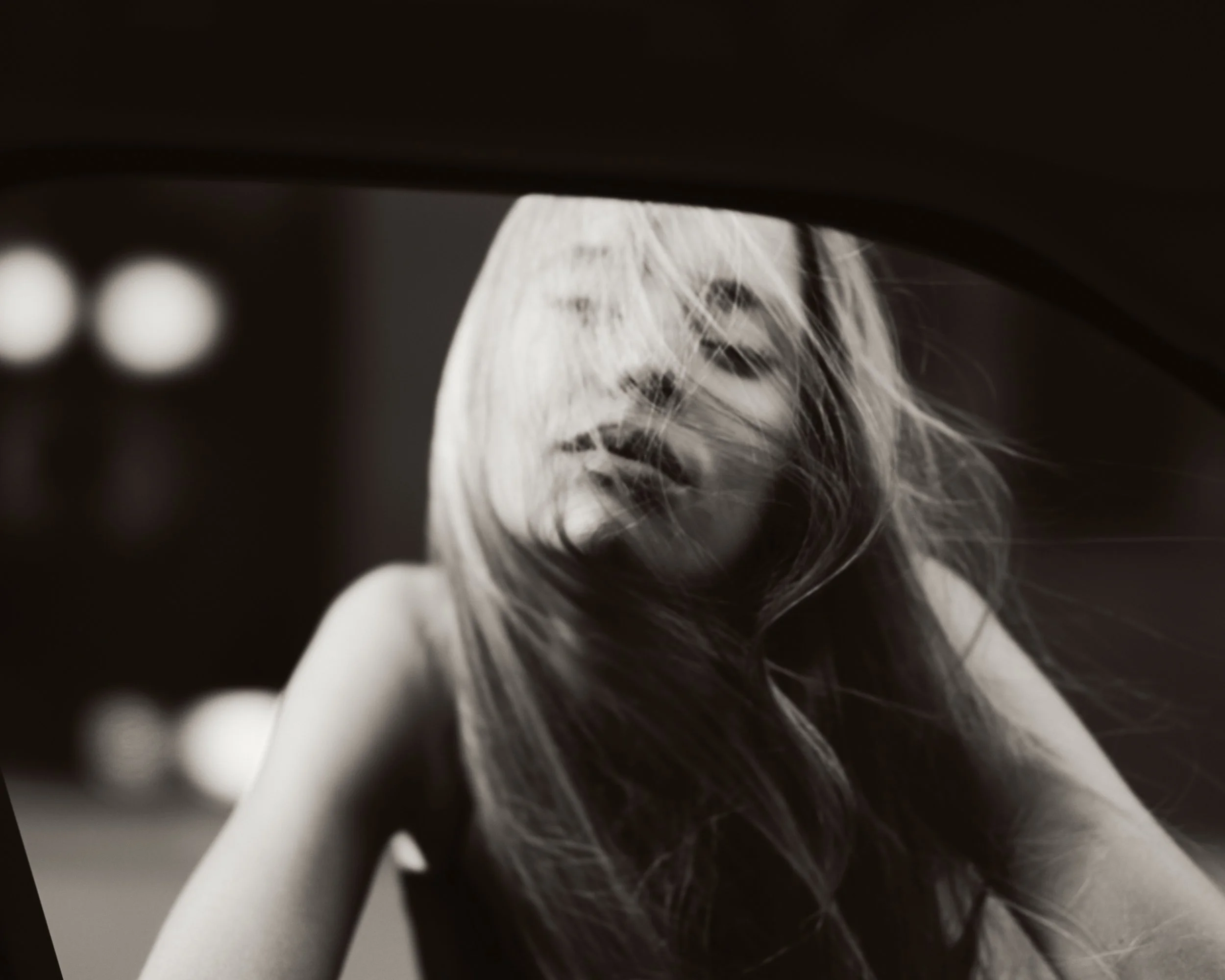 Black and white photograph of a woman with long hair covering part of her face, looking downward, with blurred lights in the background.