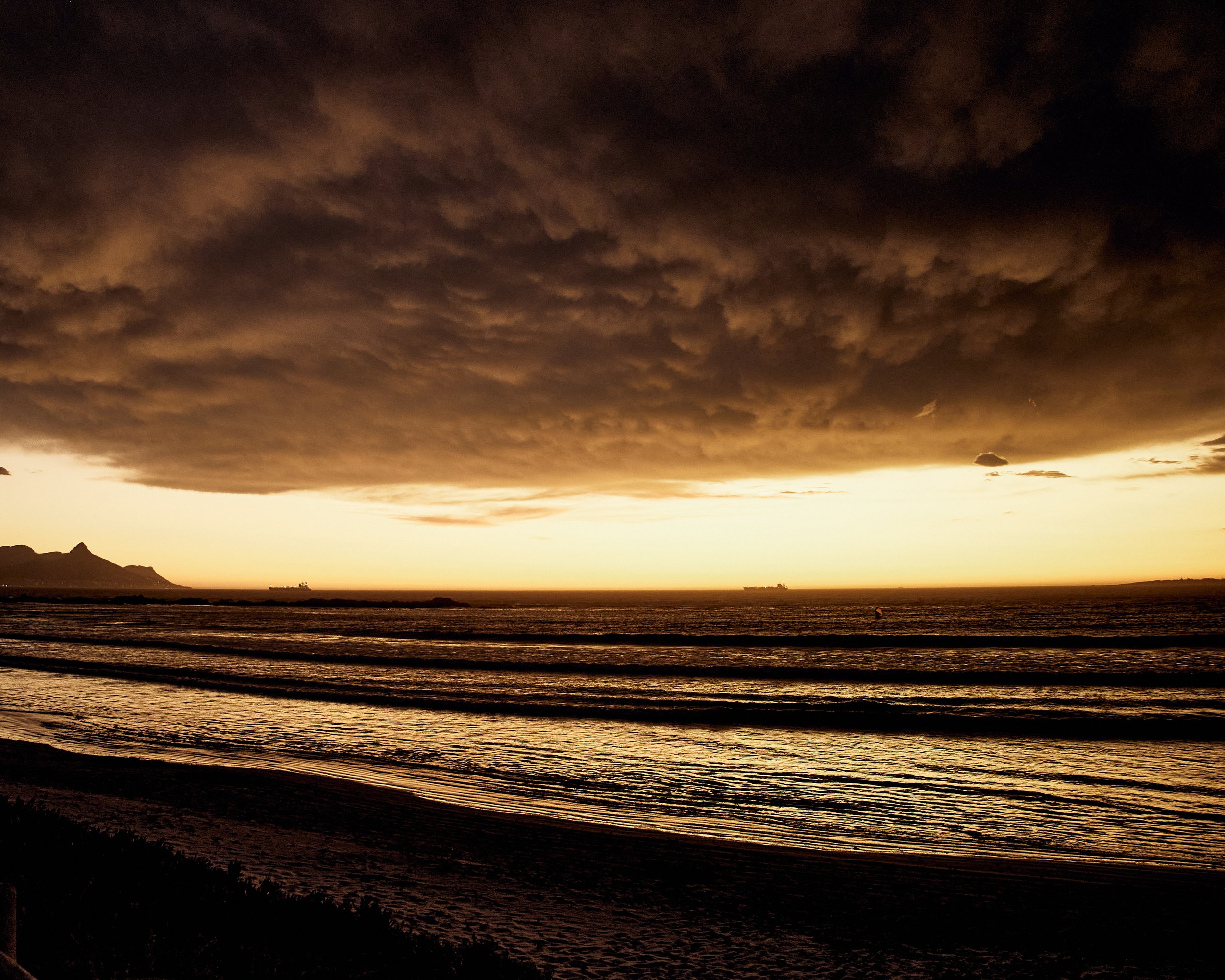 Dark, cloud-covered sky over a calm beach at sunset, with mountains and ships visible in the distance.