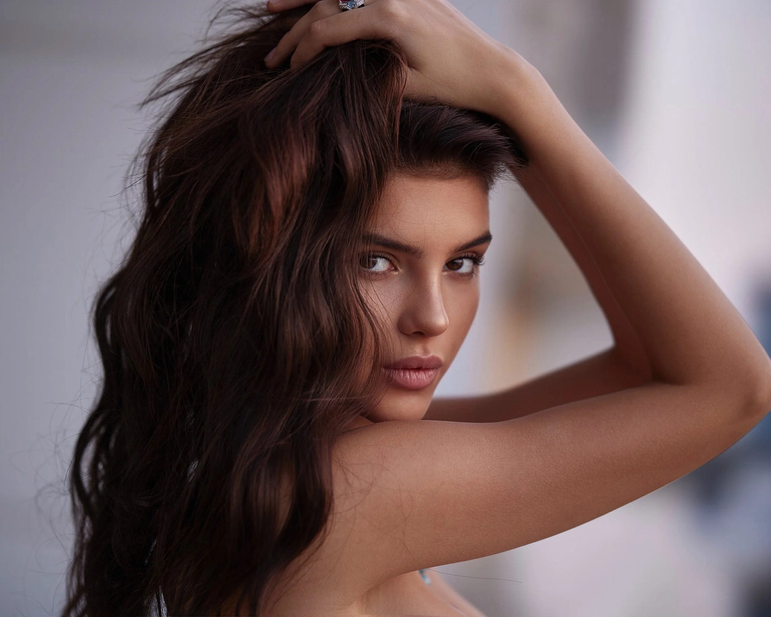 A young woman with long, wavy brown hair and light skin poses with her hand on her head, looking directly at the camera.