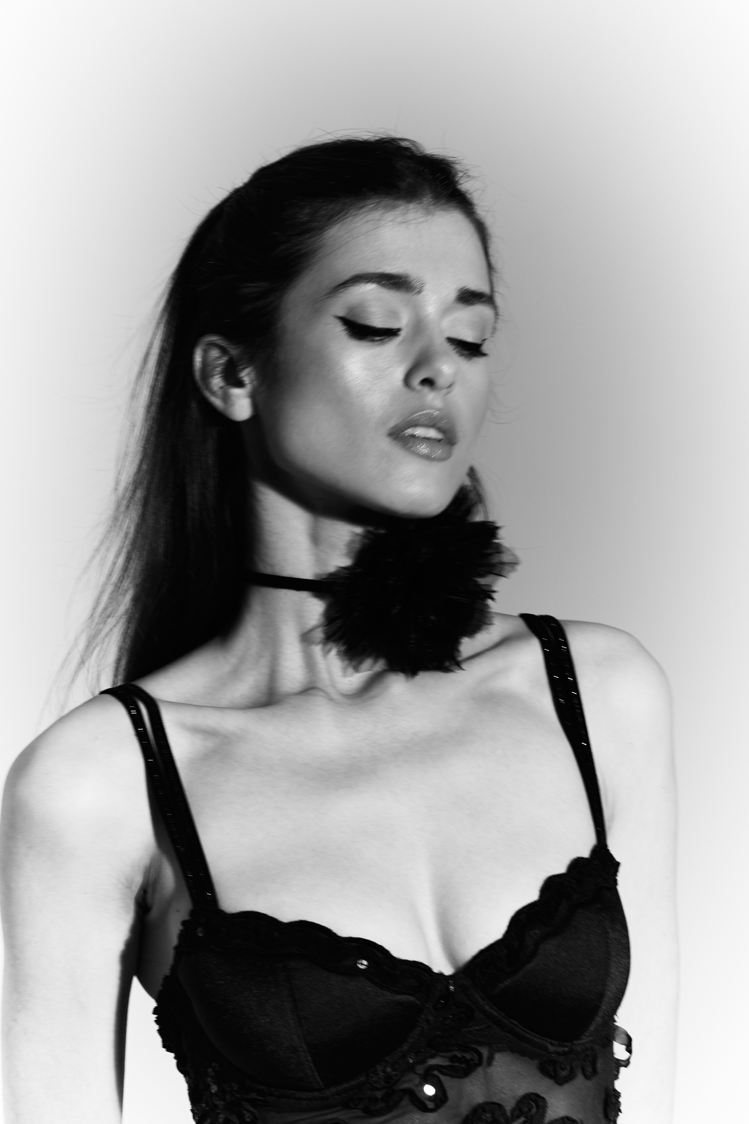 A woman with dark hair and makeup wearing lingerie and a feather choker.